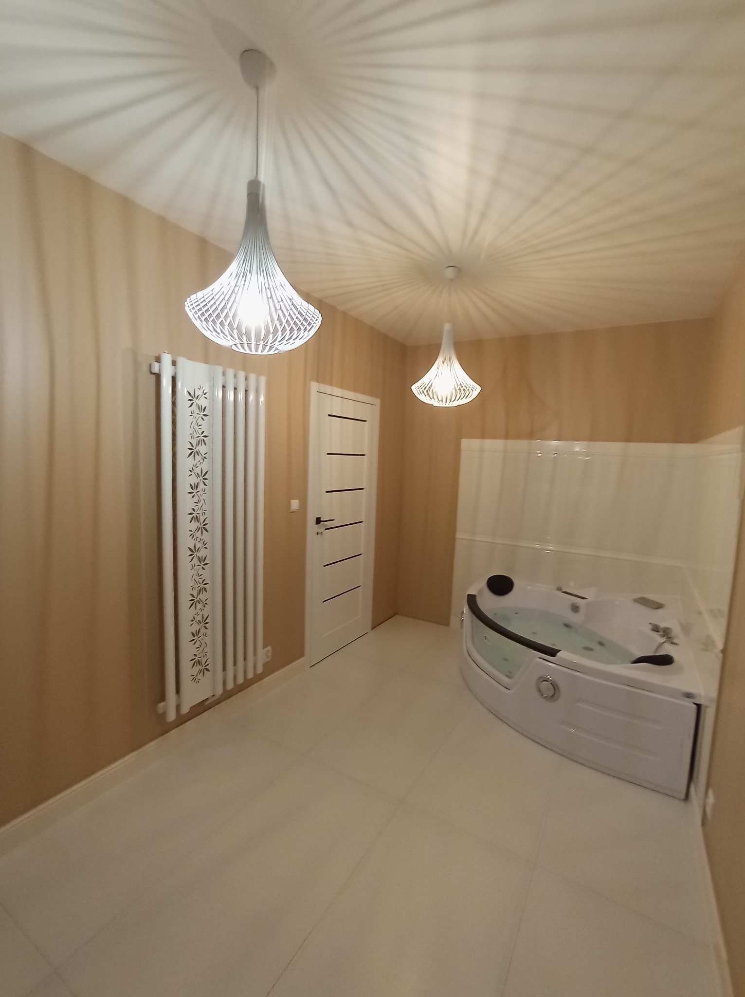 Nowoczesna łazienka z narożną wanną z hydromasażem, grzejnikiem dekoracyjnym z motywem roślinnym i designerskimi lampami wiszącymi, oświetlającymi kremowe ściany i jasną podłogę.