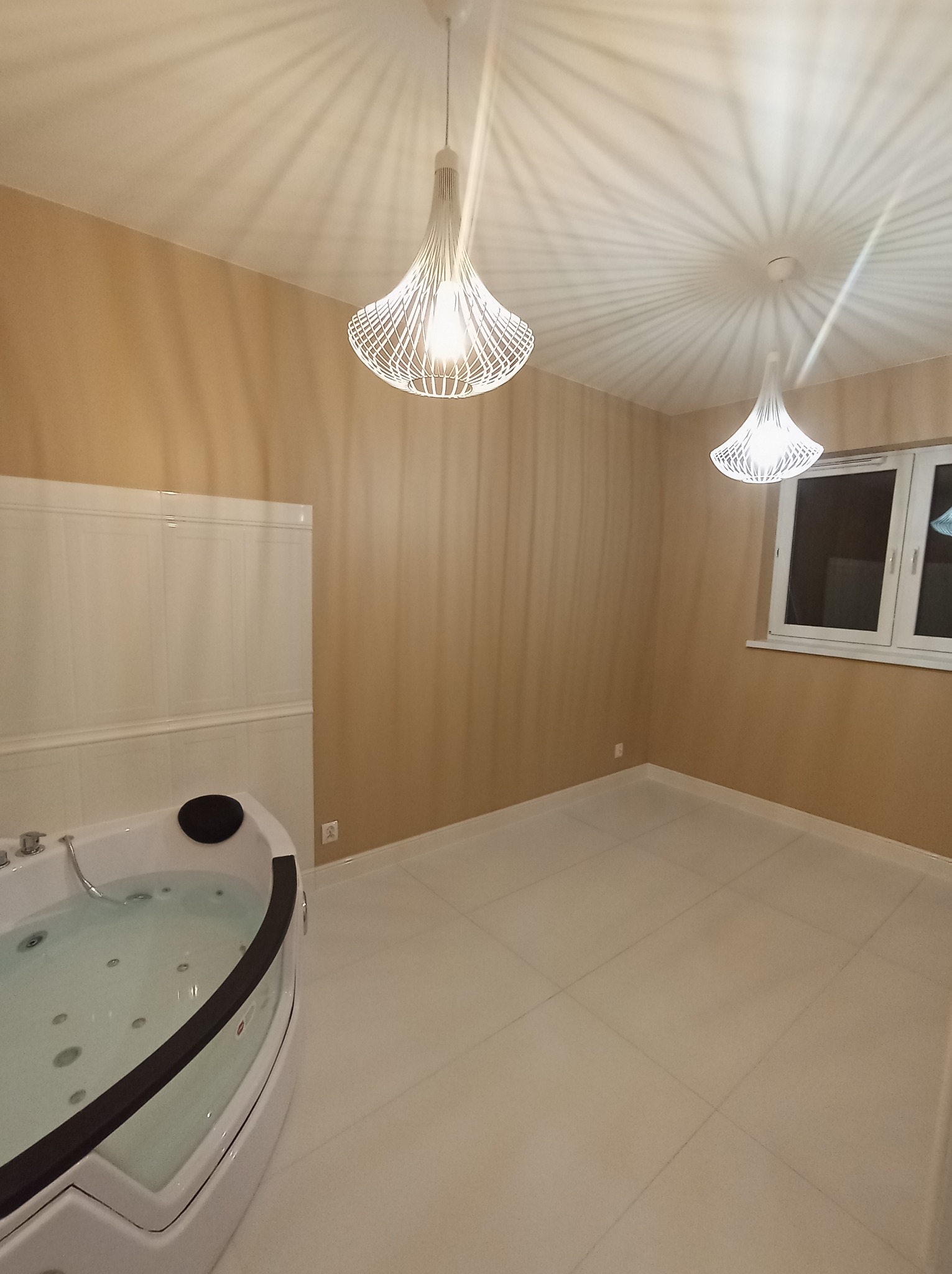 Jasna łazienka z narożną wanną jacuzzi, białymi płytkami podłogowymi i beżowymi ścianami, oświetlona dwoma nowoczesnymi lampami wiszącymi z ażurowymi kloszami.