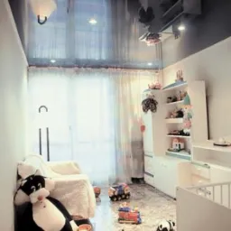 Błyszczący, ciemny sufit lustrzany w pokoju dziecięcym odbija wyposażenie: białe meble, zabawki i lampę. Widoczne okno z delikatnymi firankami. W rogu pokoju duża, pluszowa maskotka krowy.