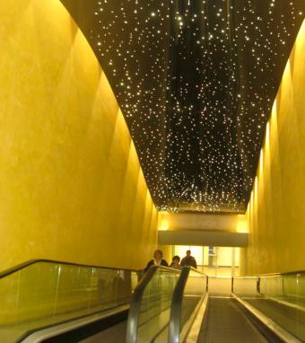 Żółte ściany korytarza z ruchomymi schodami, nad którymi znajduje się podwieszany sufit z efektem rozgwieżdżonego nieba, z widocznymi osobami w oddali.
