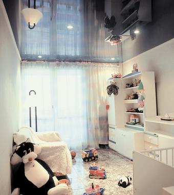 Błyszczący, ciemny sufit lustrzany w pokoju dziecięcym odbija wyposażenie: białe meble, zabawki i lampę. Widoczne okno z delikatnymi firankami. W rogu pokoju duża, pluszowa maskotka krowy.