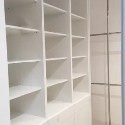 Biała, modułowa szafa wnękowa z półkami i szufladami, wyposażona w metalowy system drążków na ubrania.