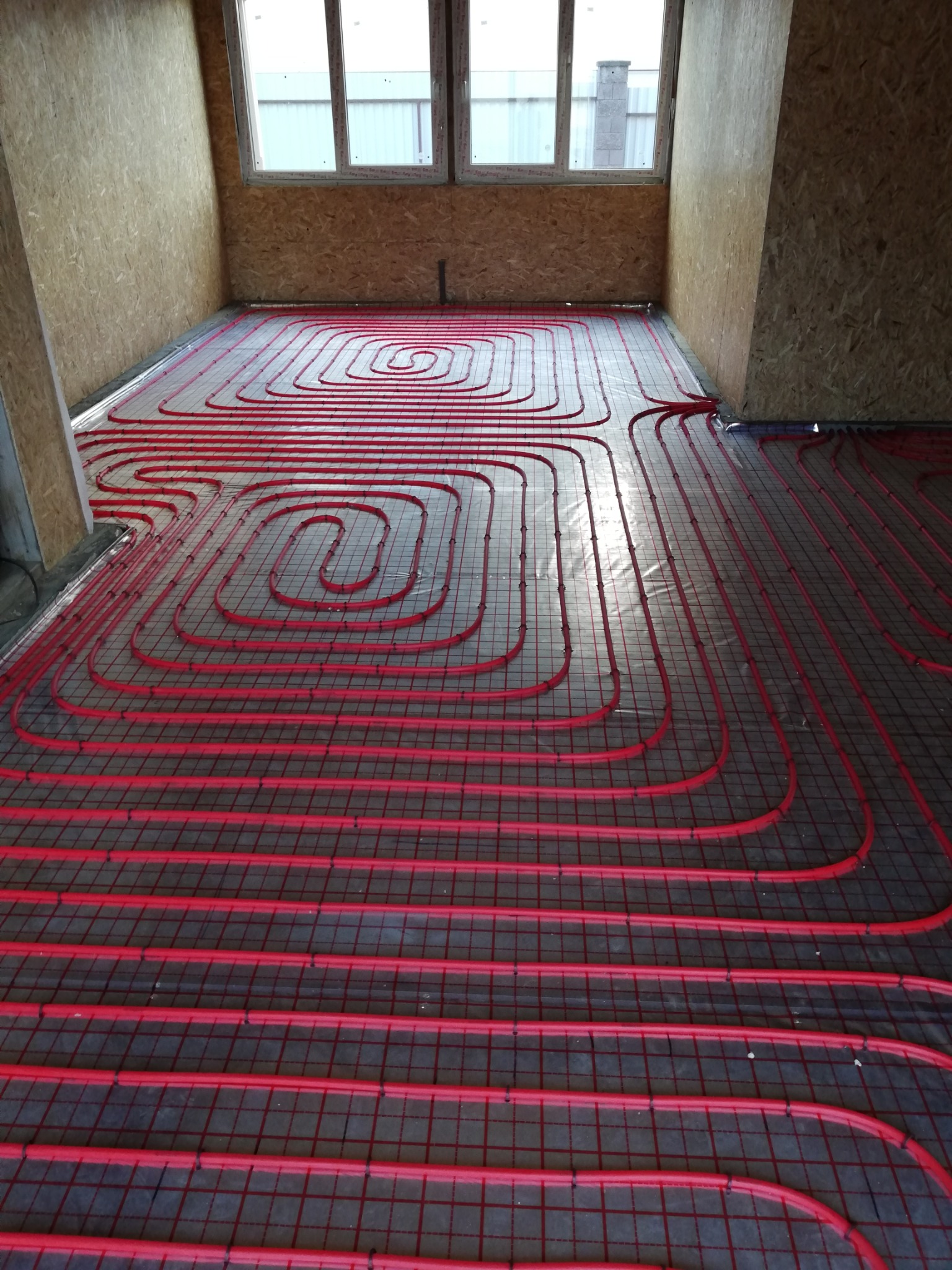 Montaż czerwonych rur ogrzewania podłogowego w nowym domu w Gdańsku, widoczne okna i surowe ściany z płyty OSB.