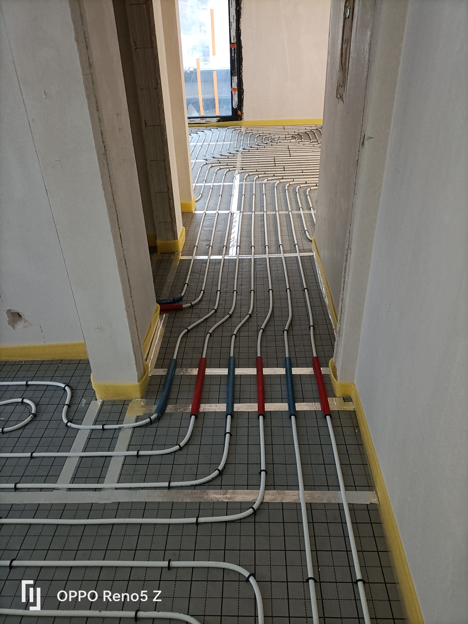 Instalacja ogrzewania podłogowego w trakcie montażu: białe rury ułożone na szarej macie z siatką, przymocowane czarnymi klipsami, widoczne izolacje przy ścianach, surowy stan wykończenia wnętrza.