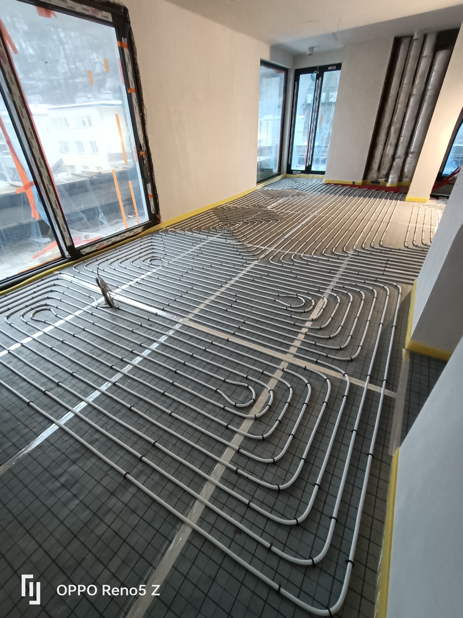 Białe rury ogrzewania podłogowego ułożone w pętle na szarej macie z siatką, widok z góry na etapie instalacji w surowym pomieszczeniu z dużymi oknami.