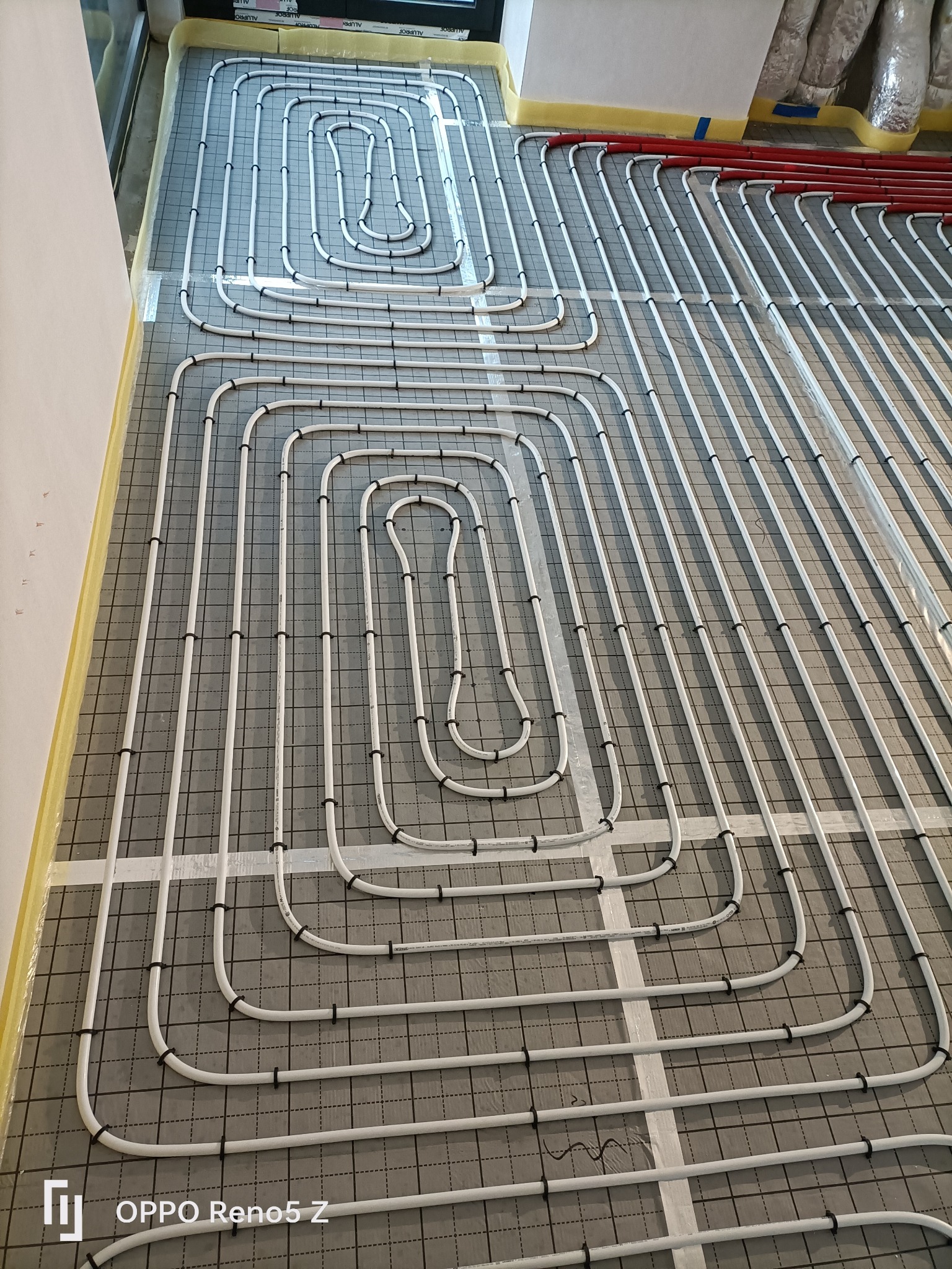 Białe rury ogrzewania podłogowego ułożone w pętle na szarej macie izolacyjnej z czarną siatką, przymocowane czarnymi klipsami, widoczne fragmenty czerwonych rur i folii ochronnej przy ścianach.