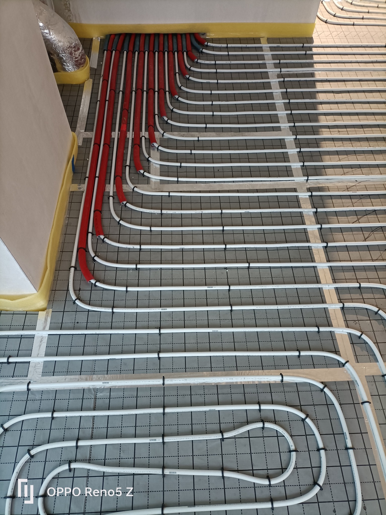 Montaż ogrzewania podłogowego: białe i czerwone rury ułożone na szarej macie z siatką, widoczne mocowania i izolacja przy ścianie.