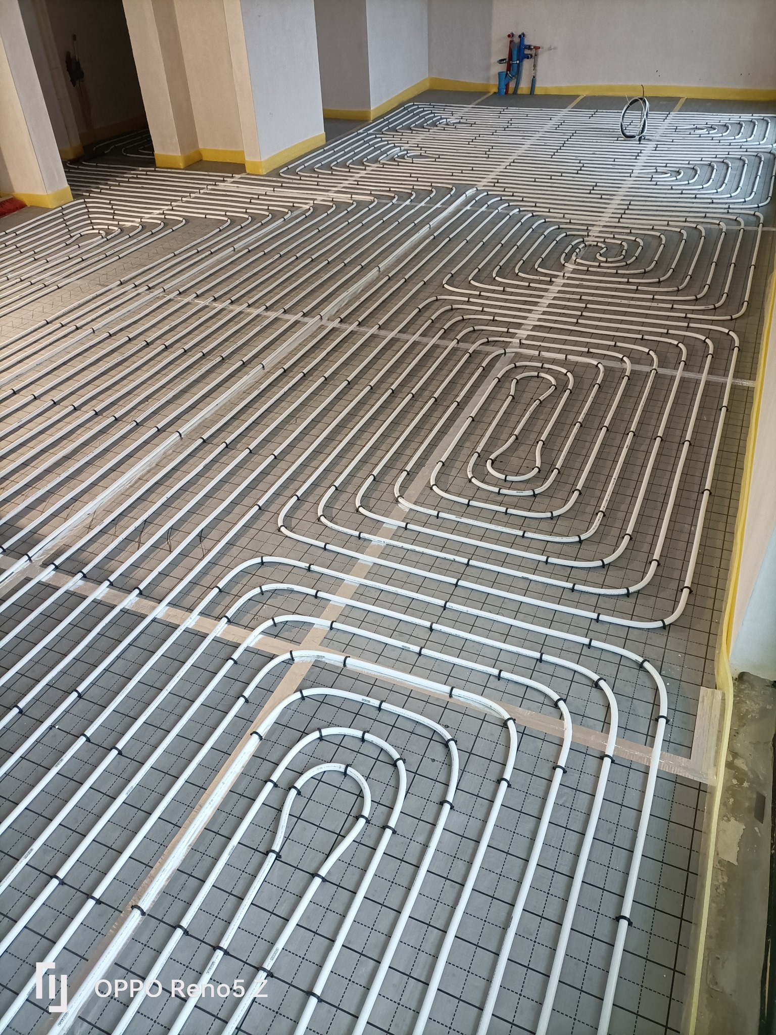 Białe rury ogrzewania podłogowego ułożone na szarej macie z siatką, widoczne elementy instalacji hydraulicznej i narzędzia w tle.