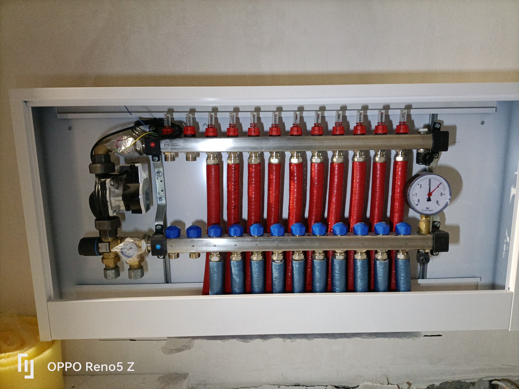Rozdzielacz ogrzewania podłogowego zamontowany w białej szafce, widoczne czerwone i niebieskie rury, manometr i zawory, instalacja w trakcie montażu.