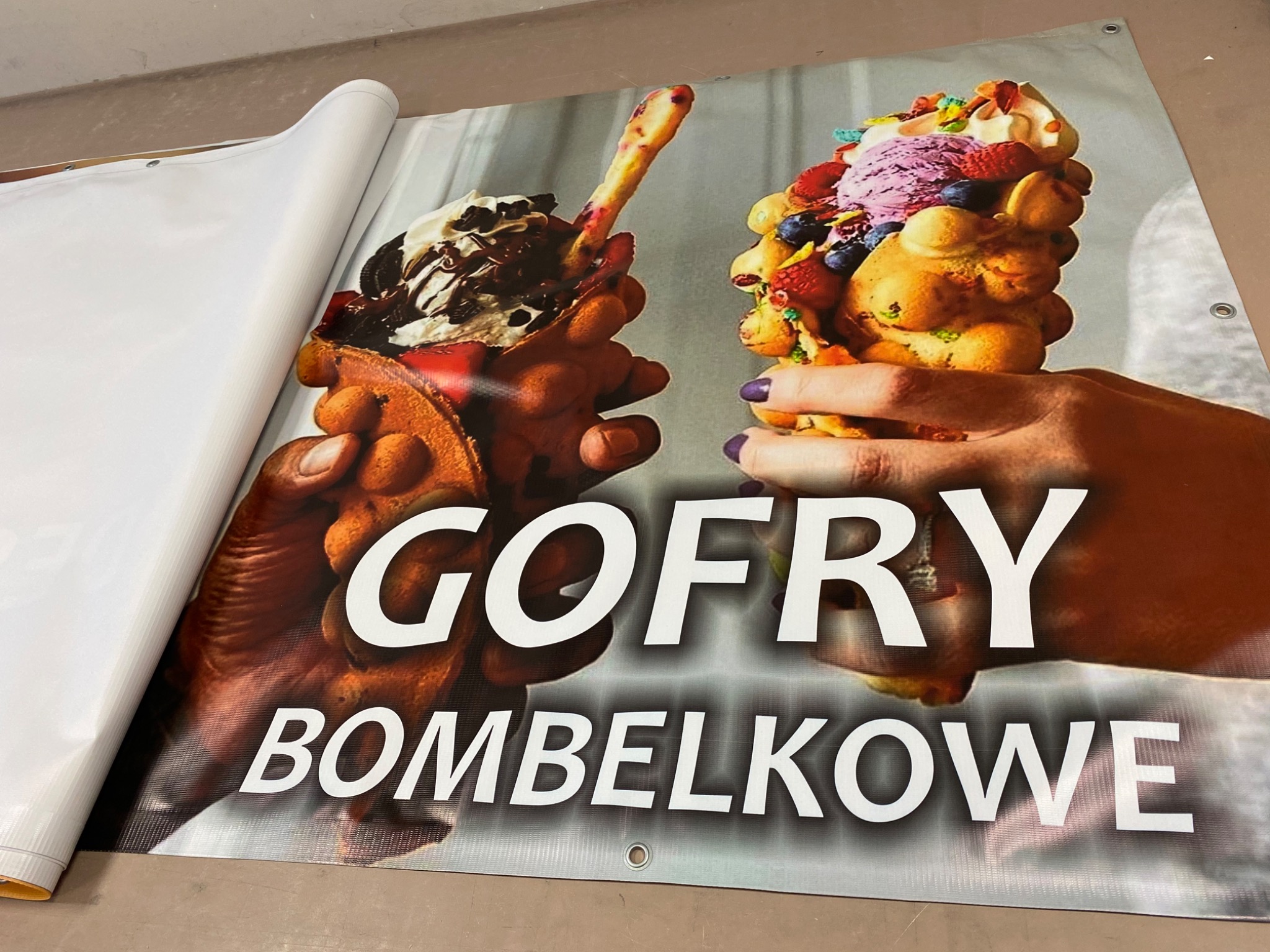 Druk wielkoformatowy baneru reklamowego z wizerunkiem gofrów bąbelkowych, częściowo zwinięty, widoczne oczka montażowe.
