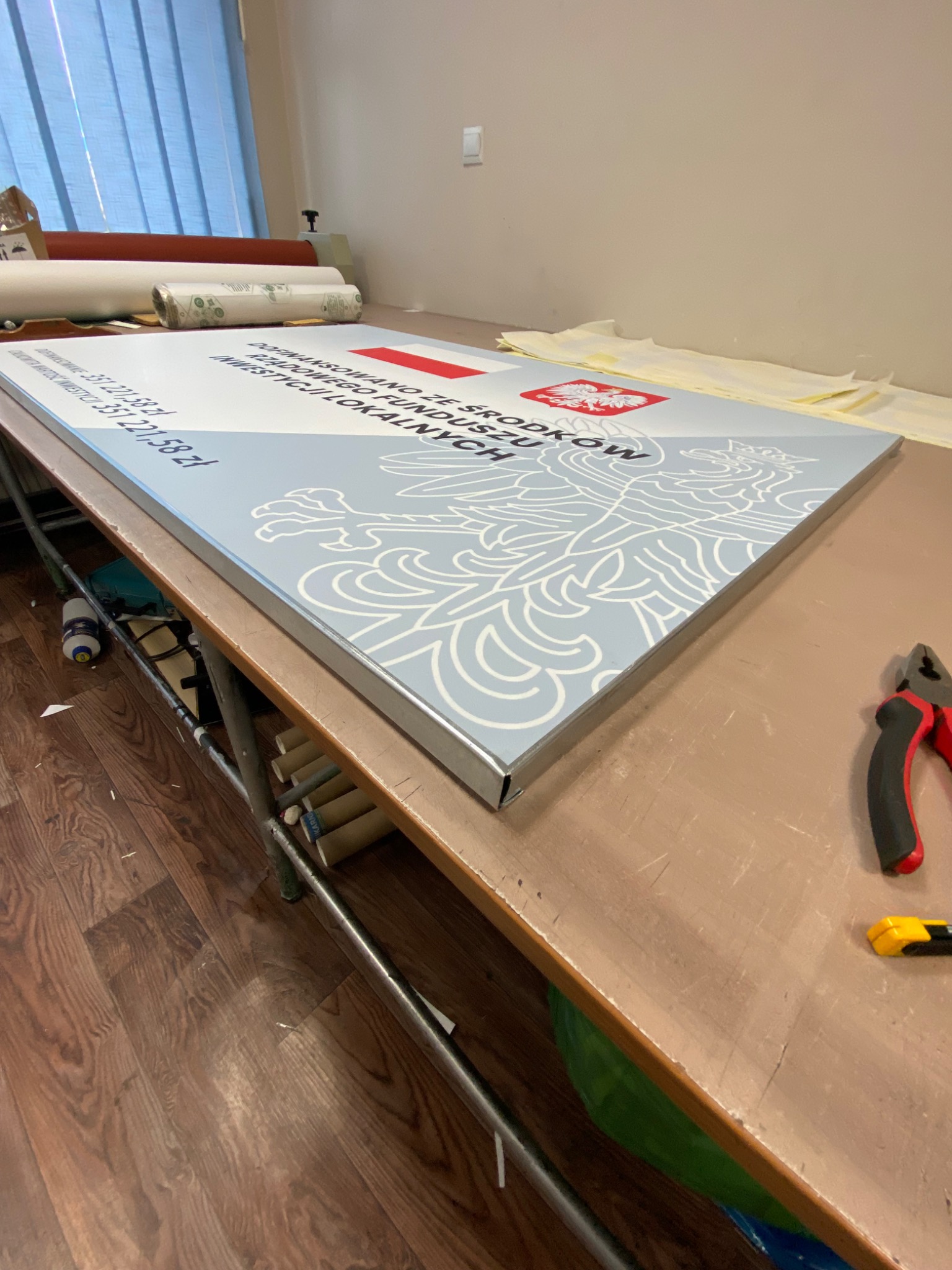 Tablica informacyjna z godłem i flagą Polski, leżąca na stole warsztatowym, obok narzędzi, w tle rolety okienne.