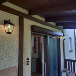 Zewnętrzna lampa w stylu retro na elewacji domu z fakturą baranka, oświetlająca wejście z przesuwnymi drzwiami i widokiem na wnętrze kuchni.