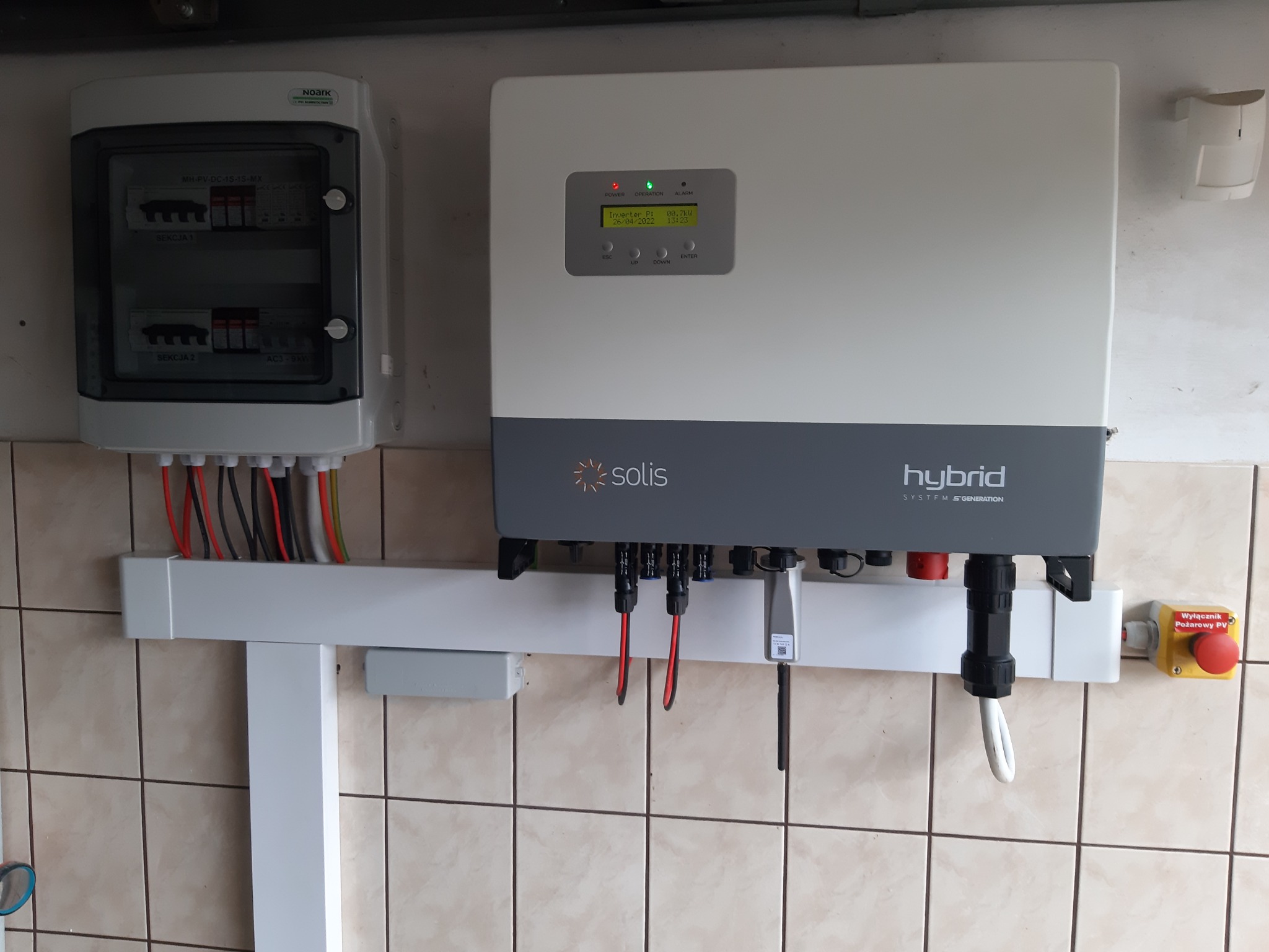 Szary inwerter hybrydowy Solis zamontowany na ścianie z beżowych płytek, obok skrzynka z bezpiecznikami i czerwony wyłącznik awaryjny.