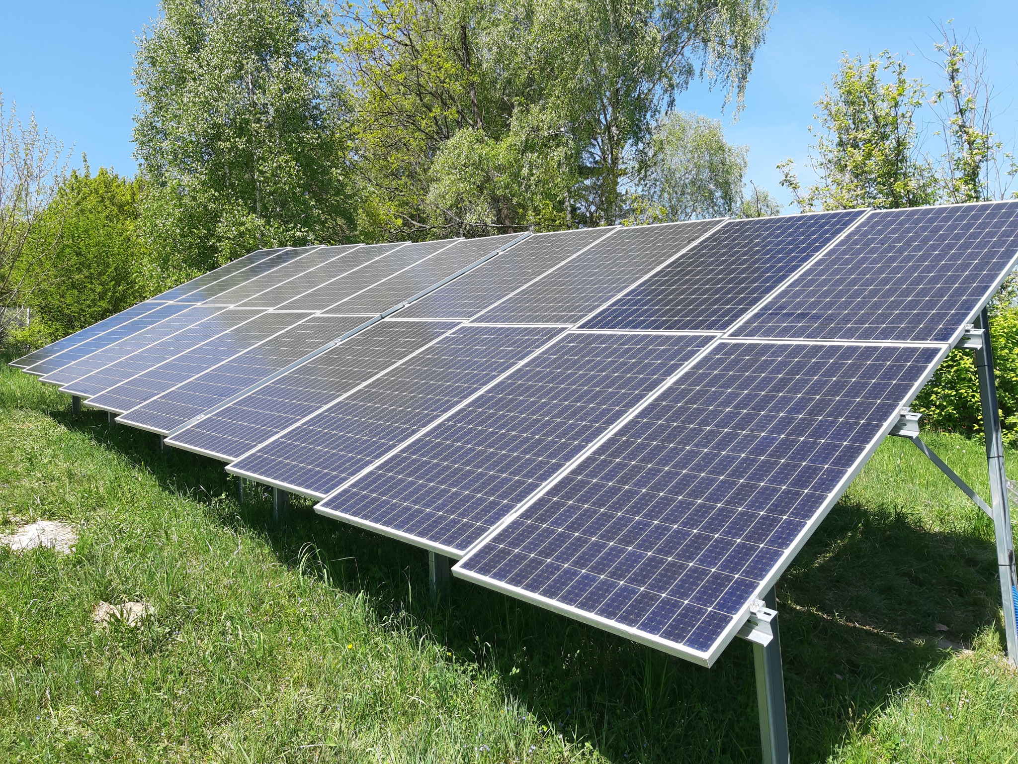 Rząd paneli fotowoltaicznych na metalowej konstrukcji, zainstalowany na trawiastym terenie z drzewami w tle, w słoneczny dzień.
