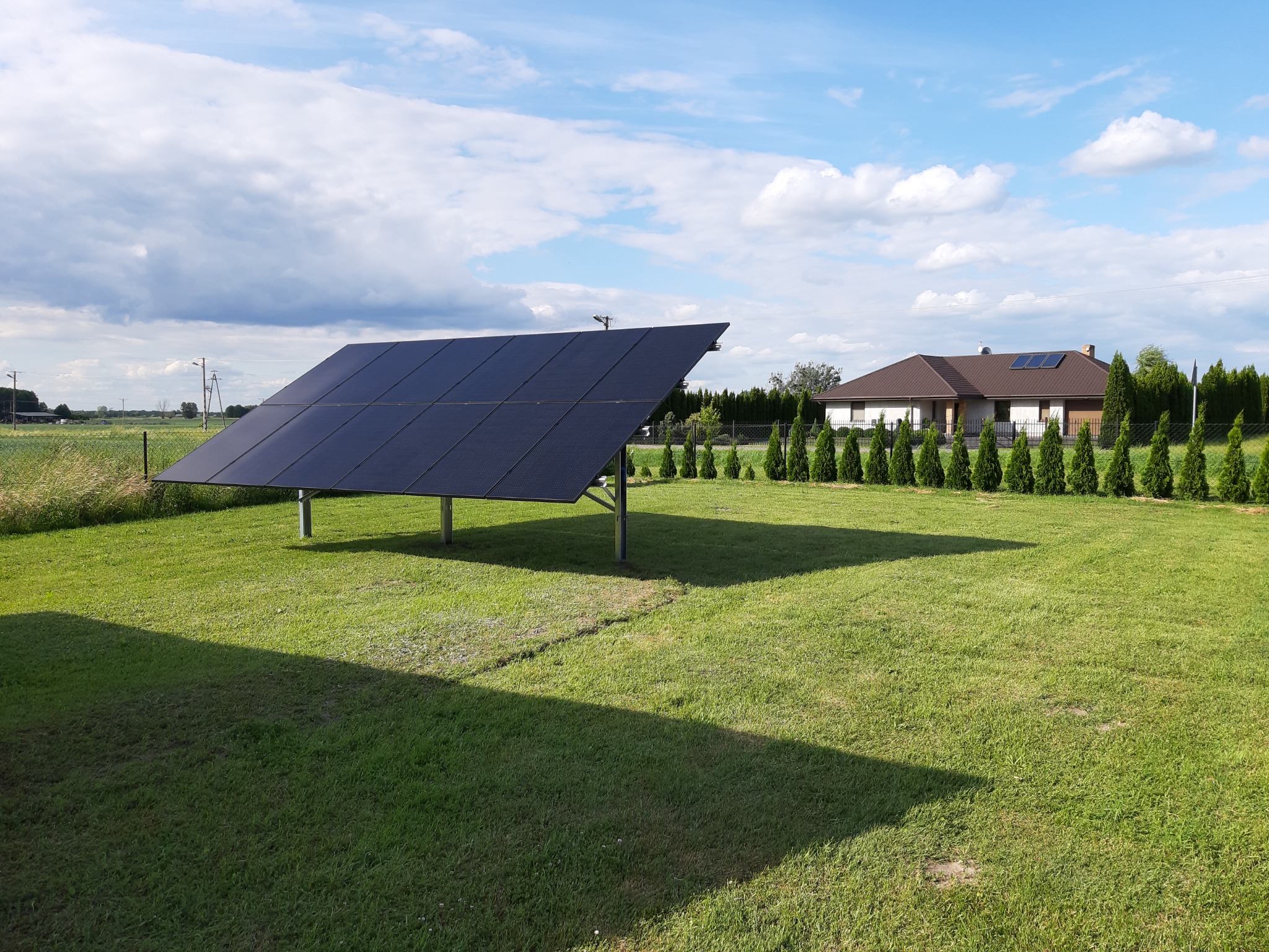 Wolnostojąca instalacja fotowoltaiczna na trawniku, w tle dom jednorodzinny i błękitne niebo z chmurami.
