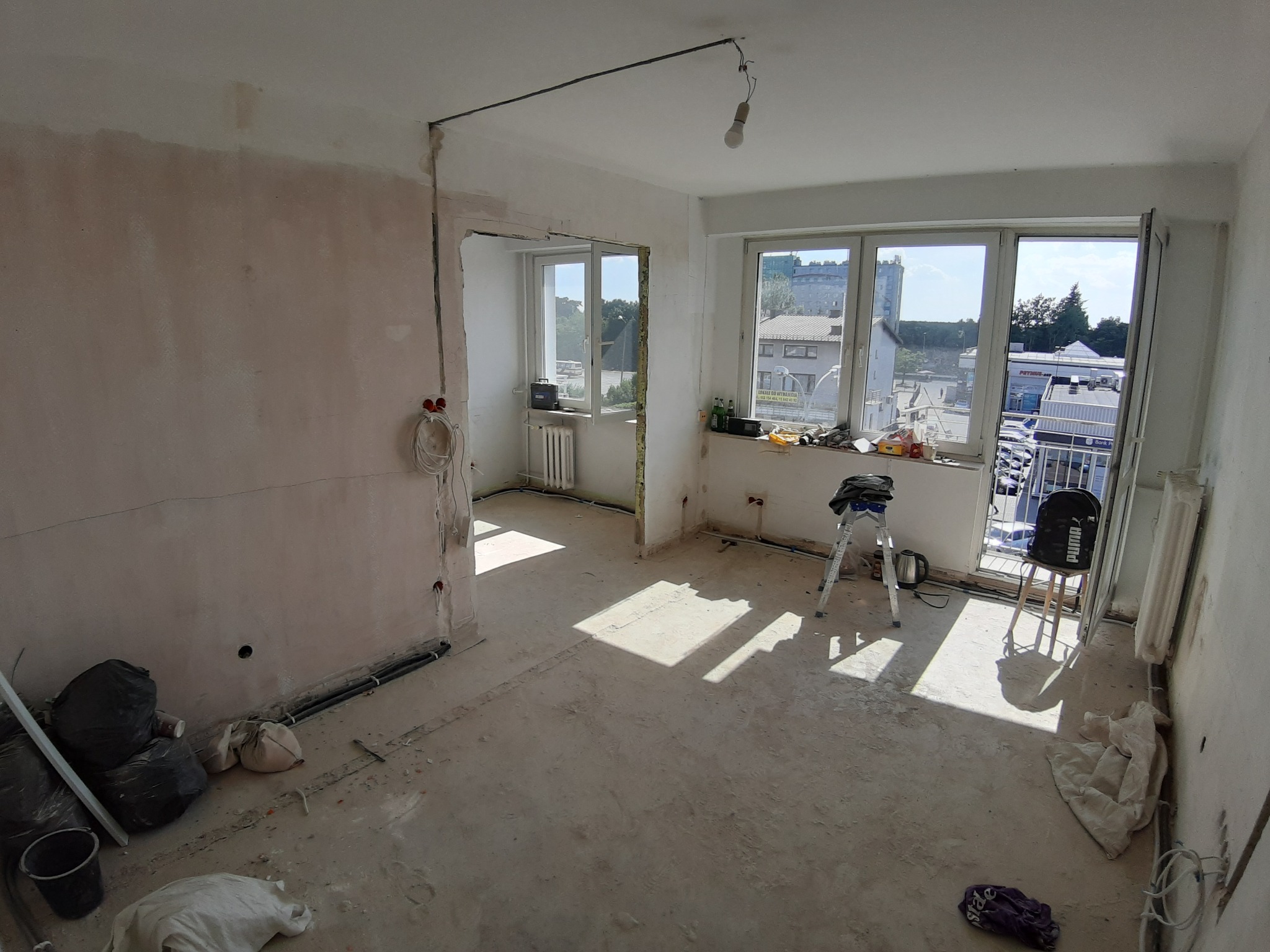 Wnętrze mieszkania w trakcie remontu, widoczne przewody elektryczne na ścianie, gołe żarówki, okna z widokiem na miasto, drabina i worki z gruzem.