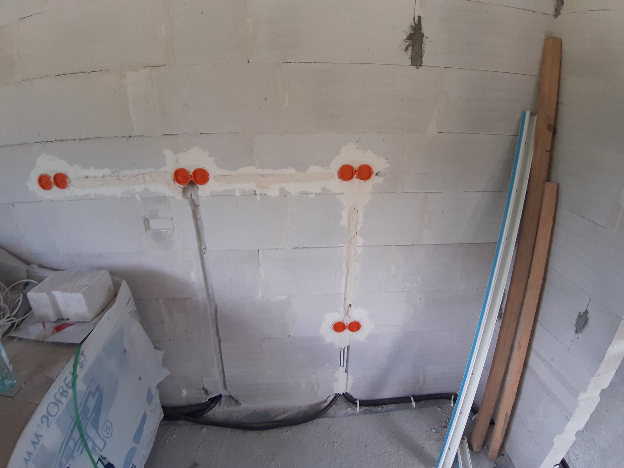 Ściana z betonu komórkowego z wyfrezowanymi bruzdami i pomarańczowymi puszkami instalacyjnymi zalewanymi gipsem, widoczne przewody elektryczne w peszlach.