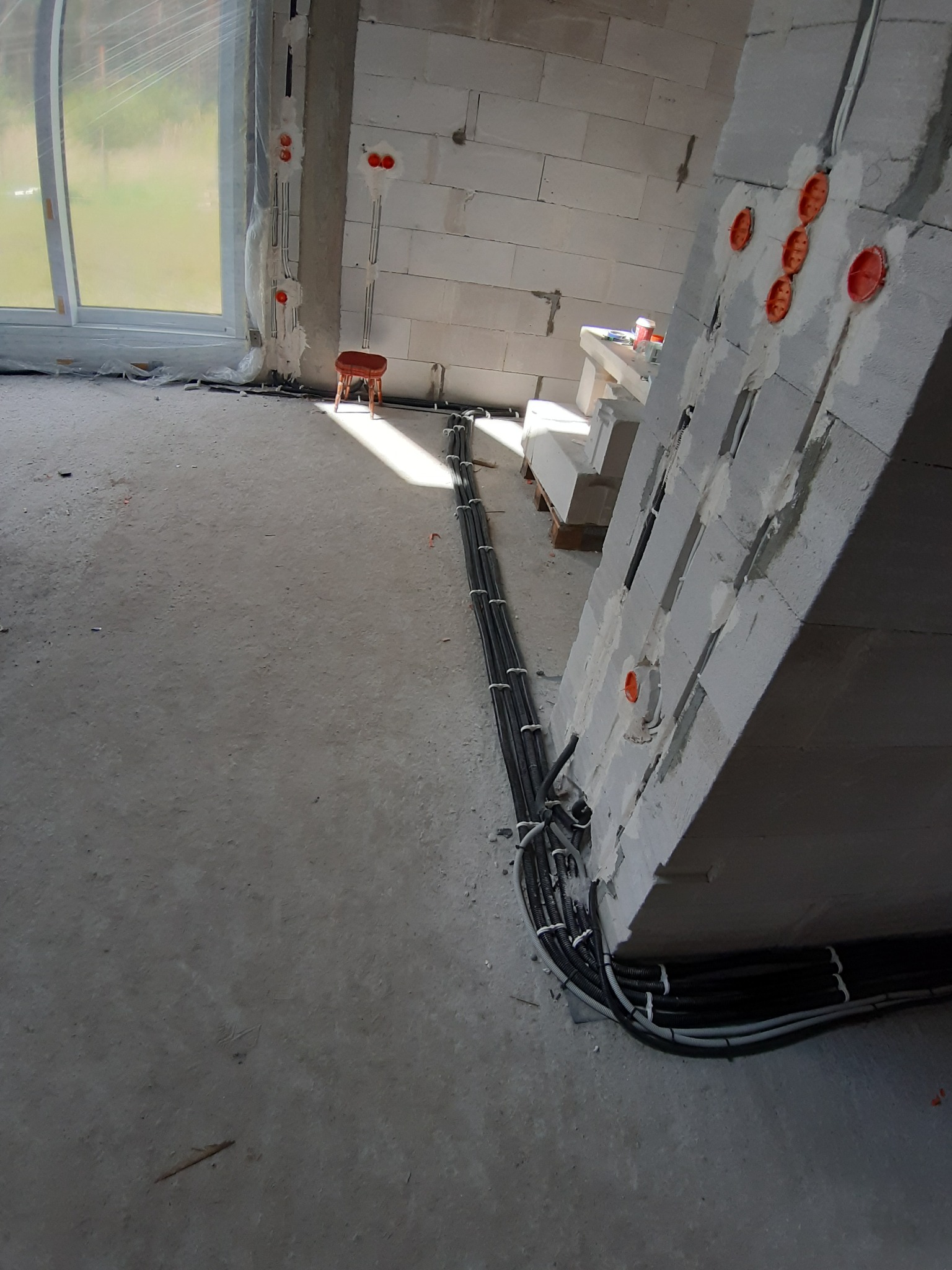 Surowe wnętrze w trakcie budowy z widocznymi przewodami elektrycznymi prowadzonymi w ścianach z betonu komórkowego i układanymi na podłodze, z pomarańczowymi puszkami instalacyjnymi w ścianach.