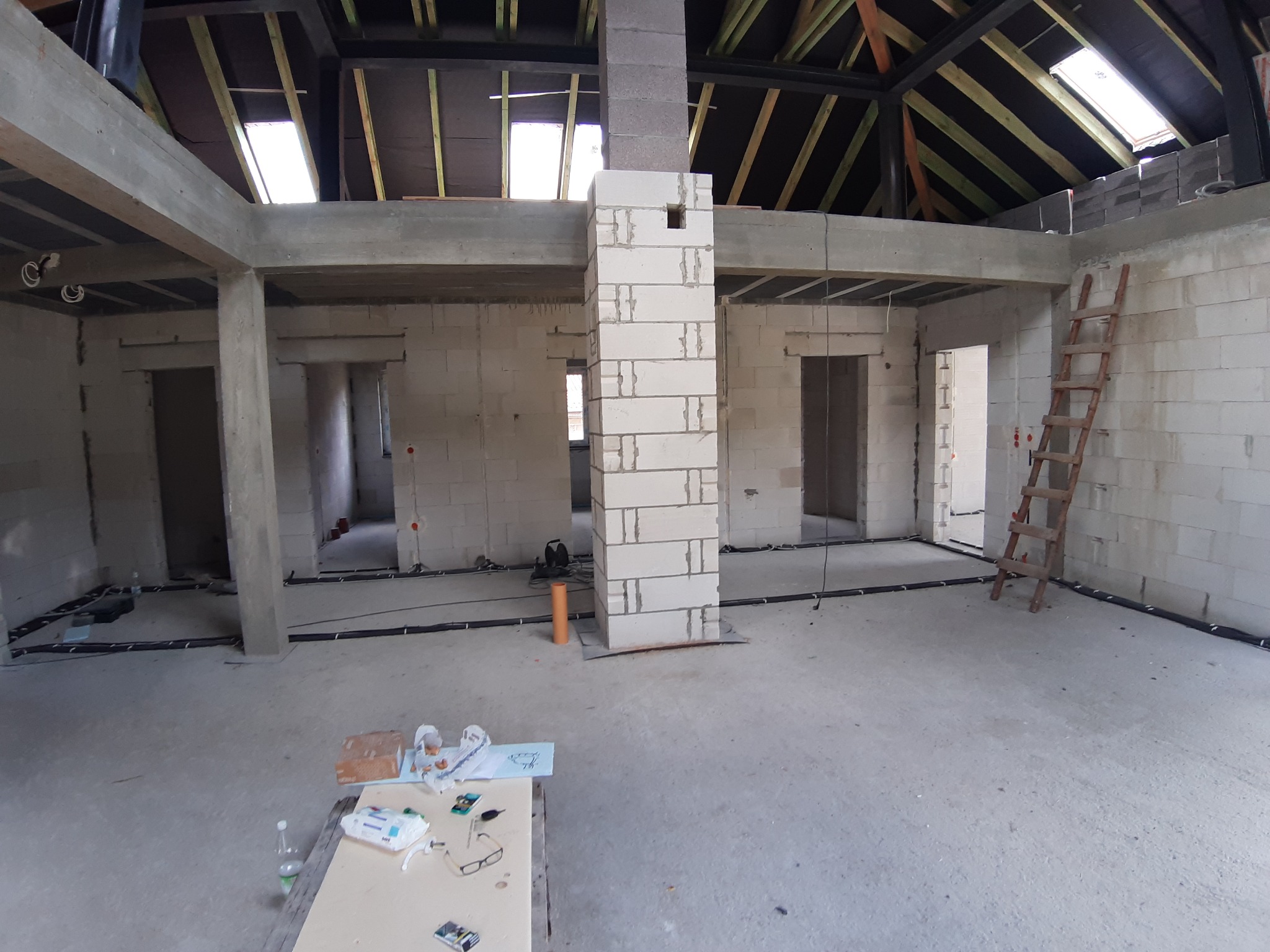 Wnętrze surowego domu w stanie deweloperskim z widoczną instalacją elektryczną w czarnych rurkach, drewnianą drabiną opartą o ścianę i stołem z narzędziami w rogu.