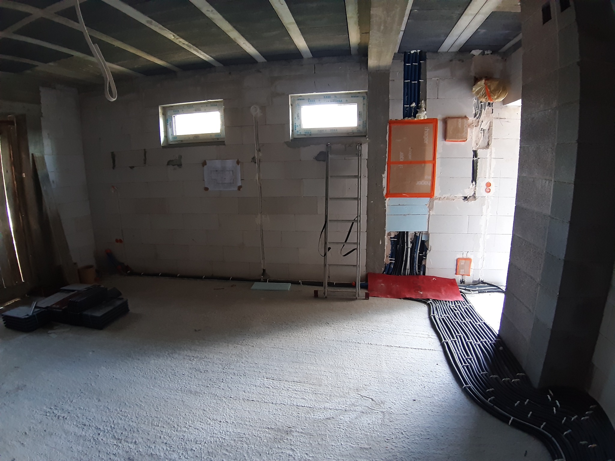 Surowy stan wnętrza budynku w trakcie instalacji elektrycznej: widoczne kable ułożone na podłodze, rury instalacyjne na ścianie, okna bez parapetów, ściany z bloczków betonowych i drabina.