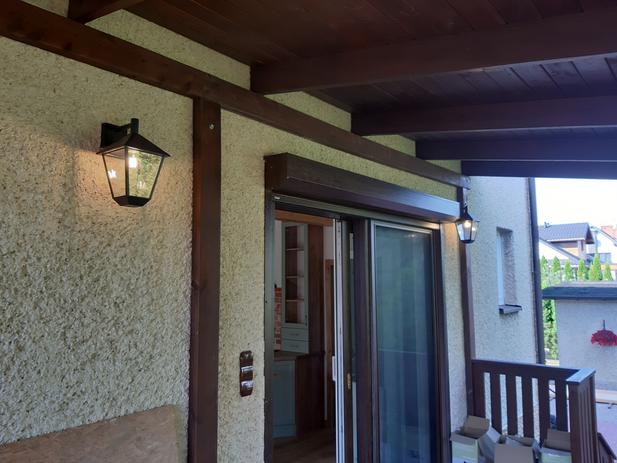 Zewnętrzna lampa w stylu retro na elewacji domu z fakturą baranka, oświetlająca wejście z przesuwnymi drzwiami i widokiem na wnętrze kuchni.