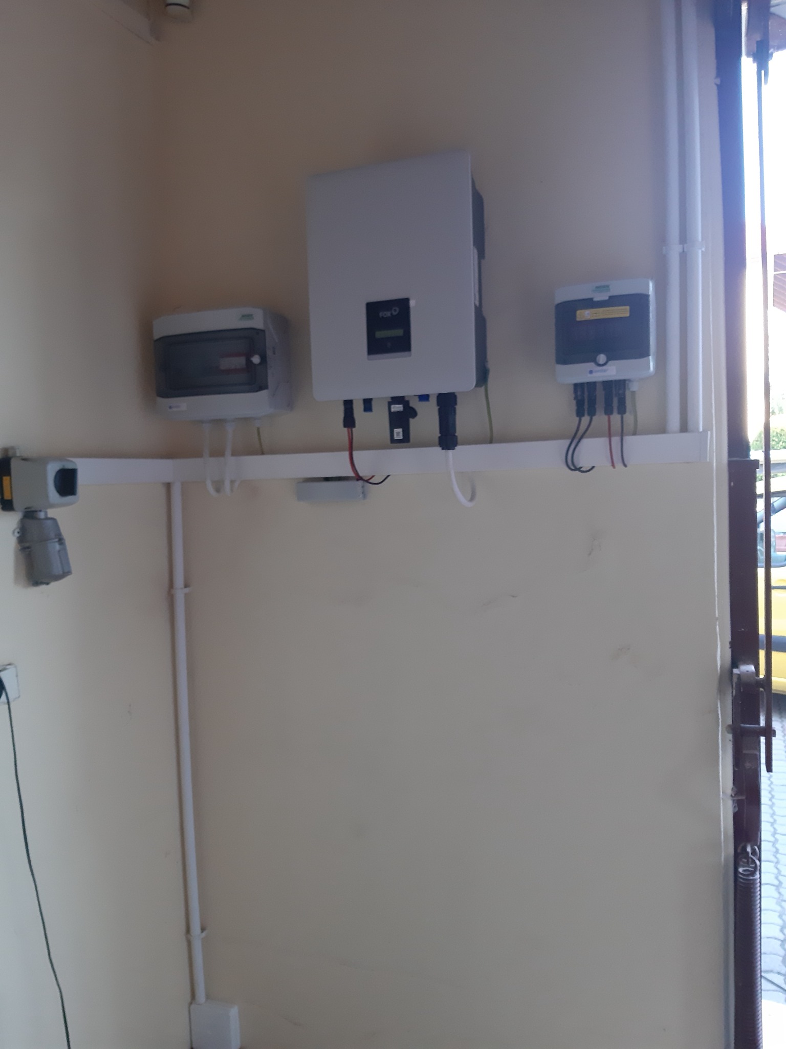 Zamontowana instalacja elektryczna z rozdzielnicą, licznikiem i inwerterem na ścianie wewnątrz budynku, widoczne przewody i elementy mocujące.