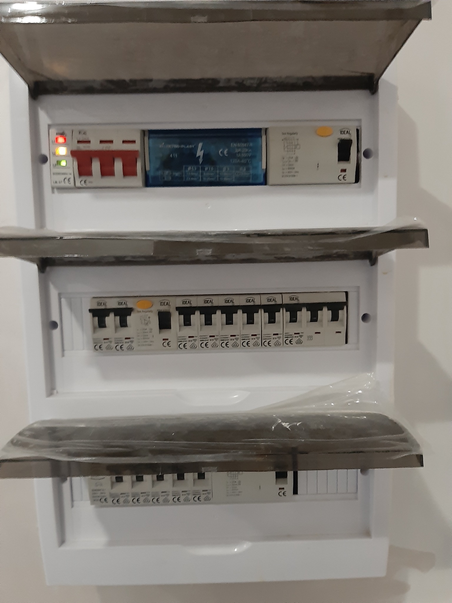 Rozdzielnica elektryczna z trzema rzędami zabezpieczeń, częściowo zakryta uchylnymi osłonami, widoczne diody sygnalizacyjne i wyłączniki różnicowoprądowe.