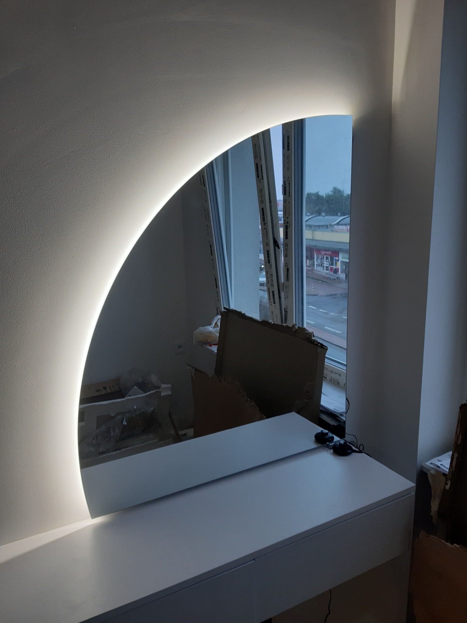 Półokrągłe lustro z podświetleniem LED zamontowane nad białą toaletką, widoczne przewody i kartony w tle, odbicie okna z widokiem na ulicę.