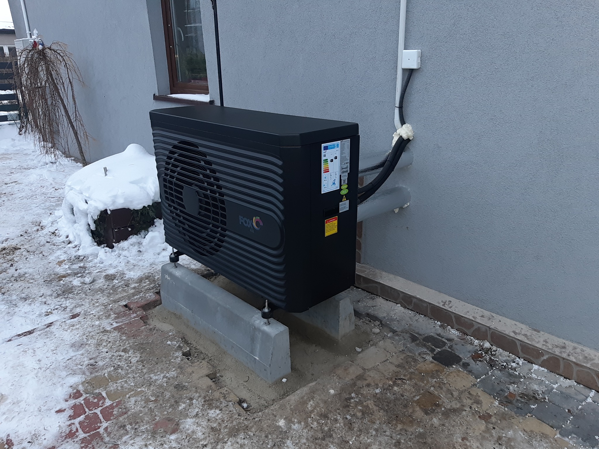 Czarna pompa ciepła marki FOX na betonowej podstawie przy szarej ścianie budynku w zimowej scenerii.