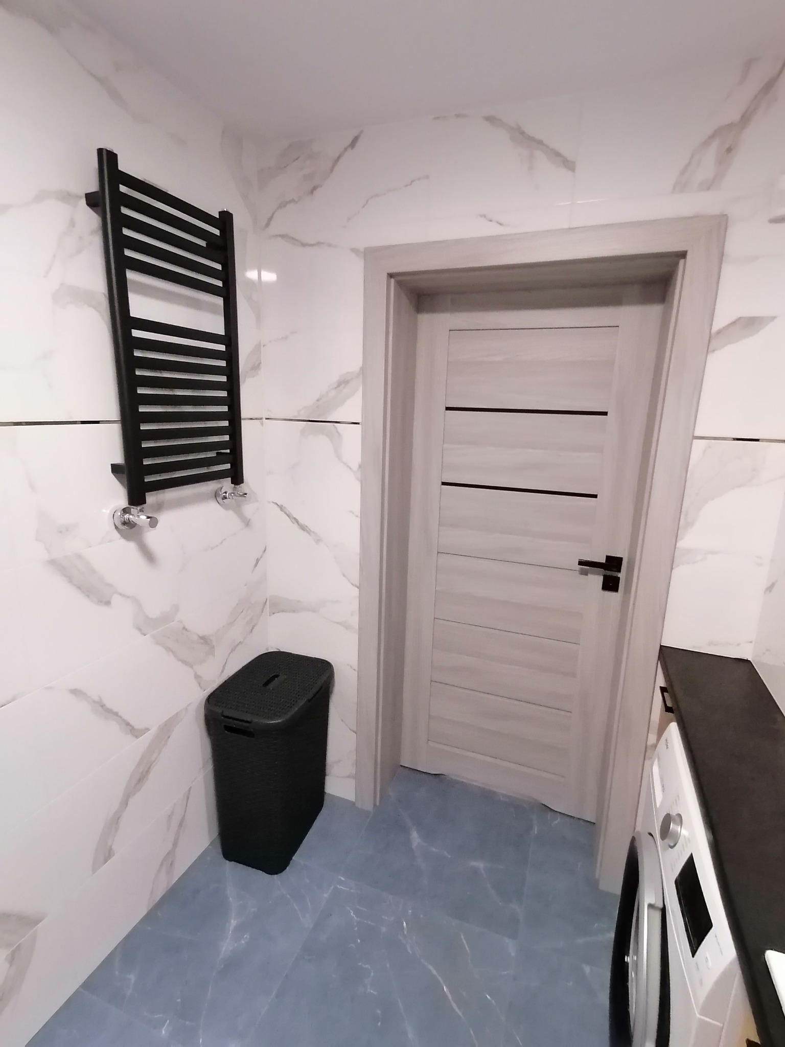 Nowoczesna łazienka z marmurowymi płytkami, czarnym grzejnikiem drabinkowym, koszem na pranie i jasnymi drzwiami z poziomymi paskami.