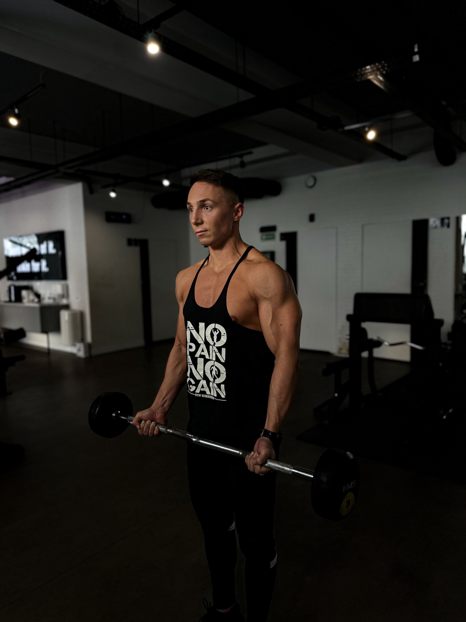Mężczyzna w czarnej koszulce z napisem 'NO PAIN NO GAIN' podnosi sztangę w siłowni, patrząc w lewo.