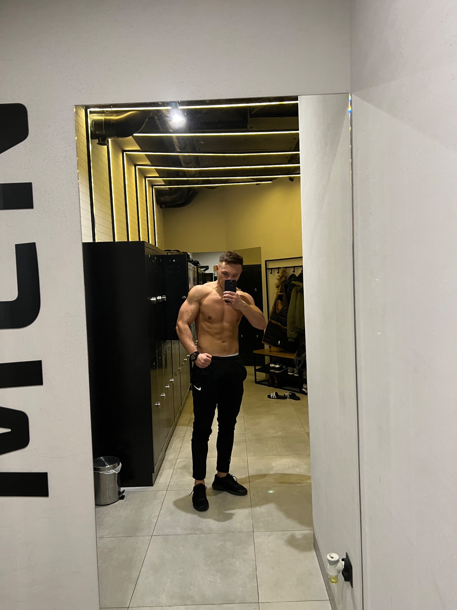 Mężczyzna bez koszulki pozuje do zdjęcia w lustrze na tle szatni siłowni z szafkami i wieszakami na ubrania.