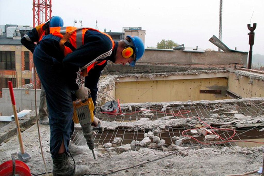 Pracownik w kasku i kamizelce używa młota pneumatycznego do rozbijania betonowej konstrukcji zbrojonej siatką, widoczne gruz i fragmenty budynku w tle.