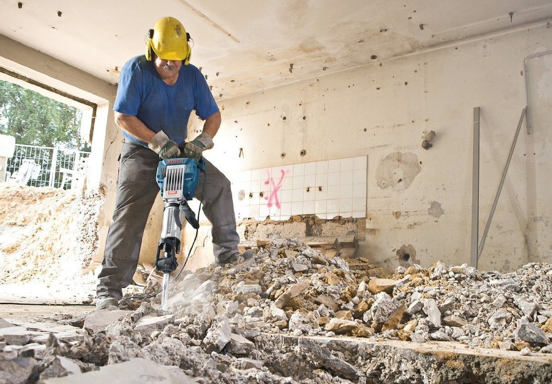 Mężczyzna w kasku i ochronnikach słuchu używa młota pneumatycznego Bosch do rozbijania podłogi w pomieszczeniu z częściowo usuniętymi płytkami ściennymi i widocznymi otworami w ścianach,...