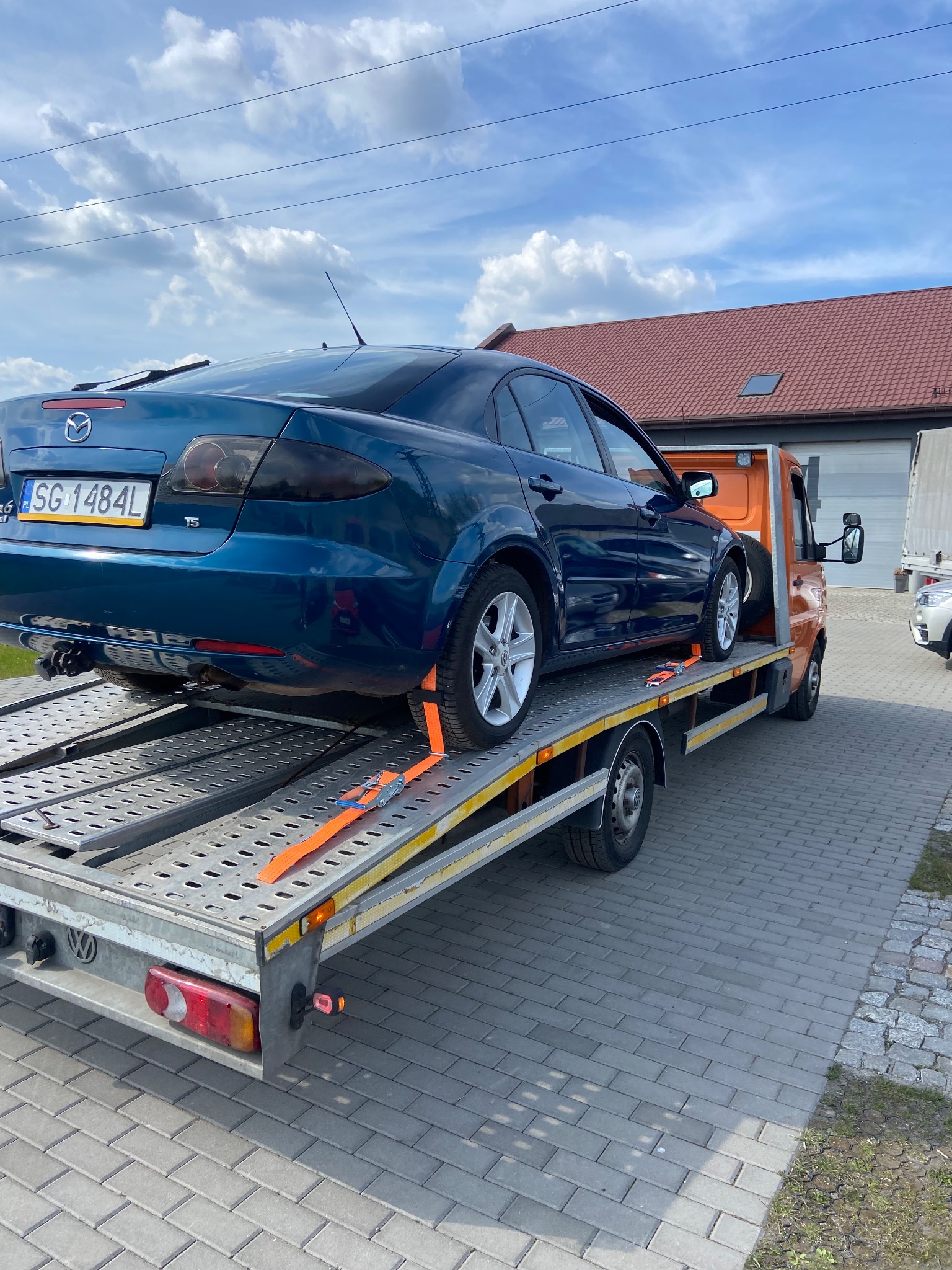 Niebieska Mazda 6 zabezpieczona pomarańczowymi pasami na srebrnej platformie lawety, widok z tyłu na tle budynku z czerwoną dachówką i błękitnego nieba.