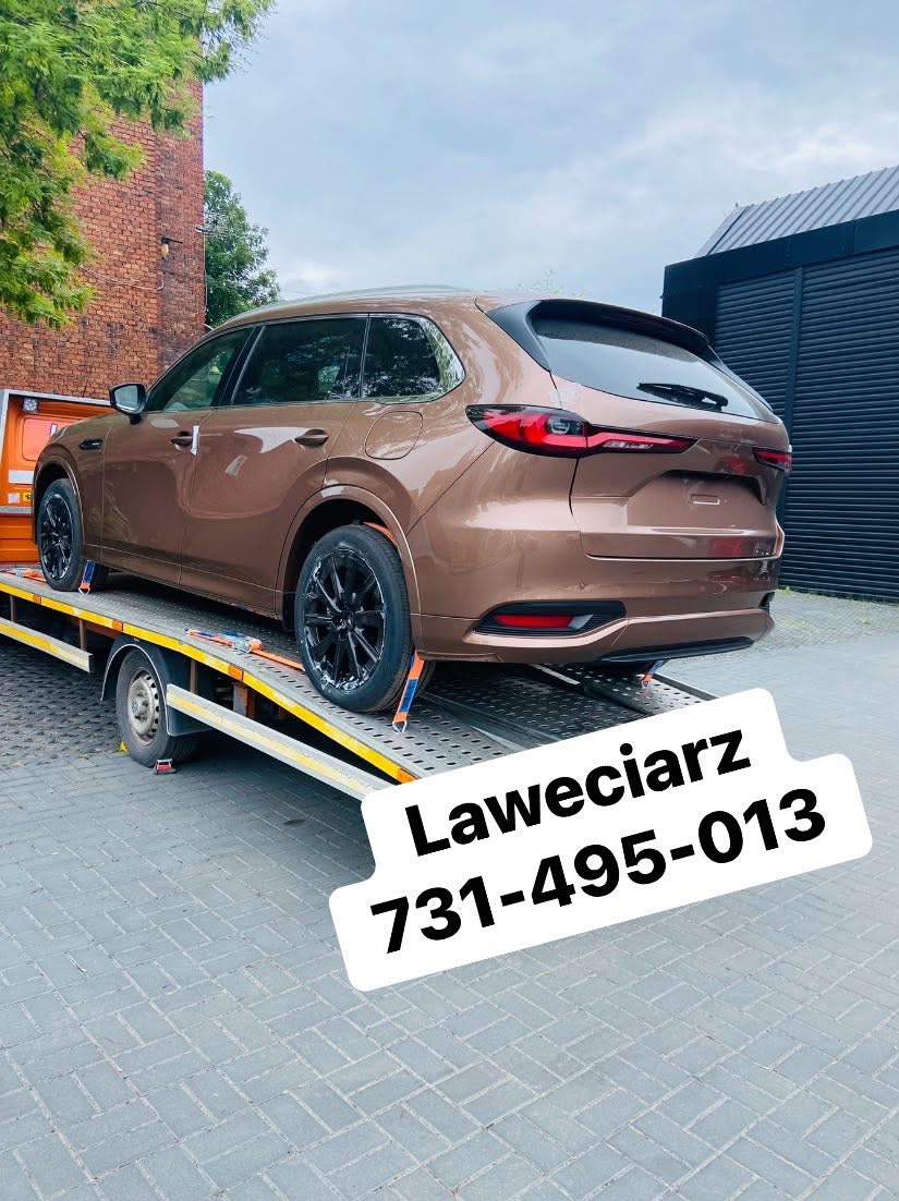 Brązowe auto typu kombi na żółtej lawecie, zabezpieczone pasami transportowymi. Widok z tyłu, ceglany budynek w tle.