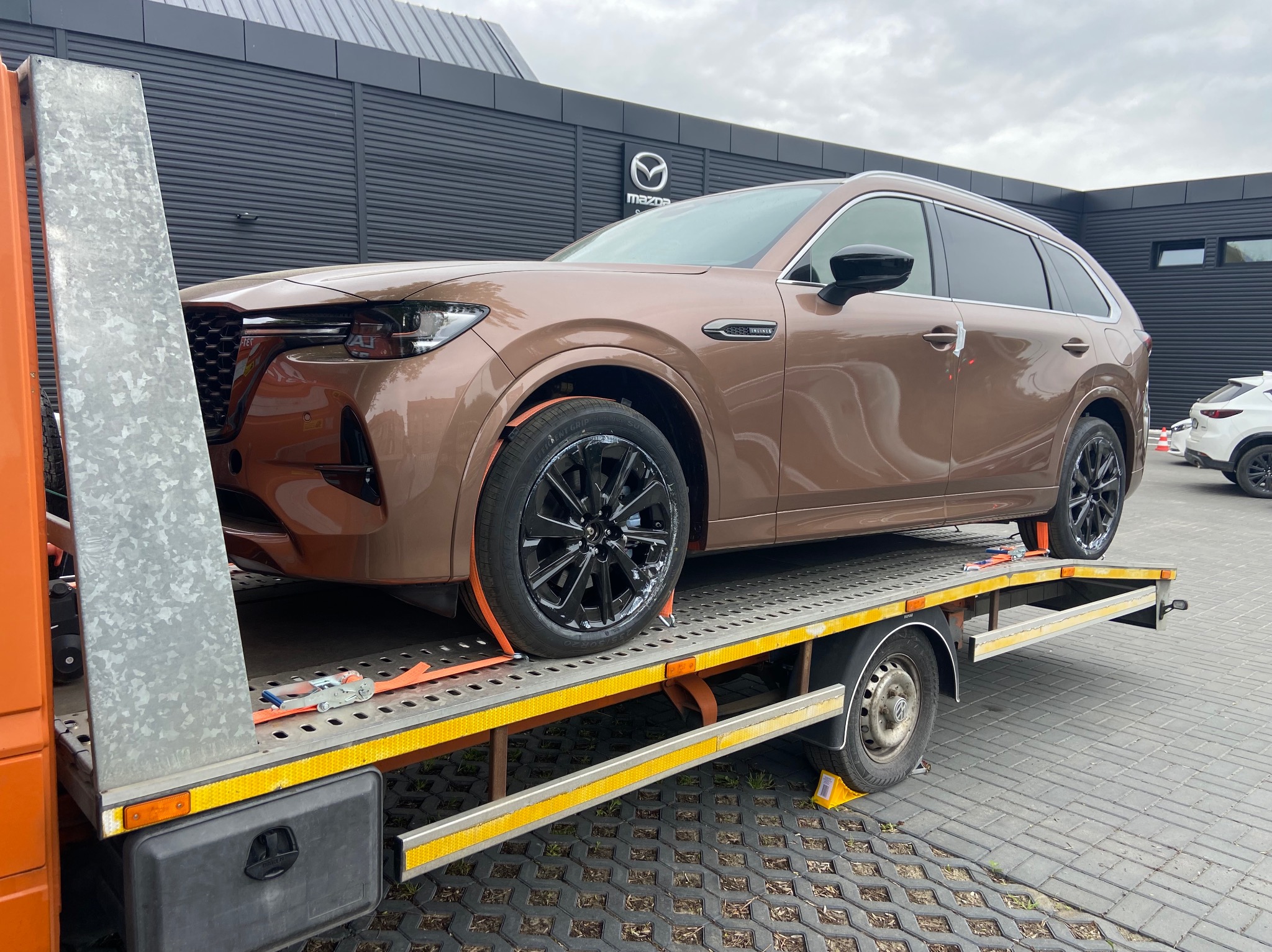 Brązowa Mazda CX-90 na platformie lawety, zabezpieczona pomarańczowymi pasami transportowymi, na tle salonu samochodowego Mazda.