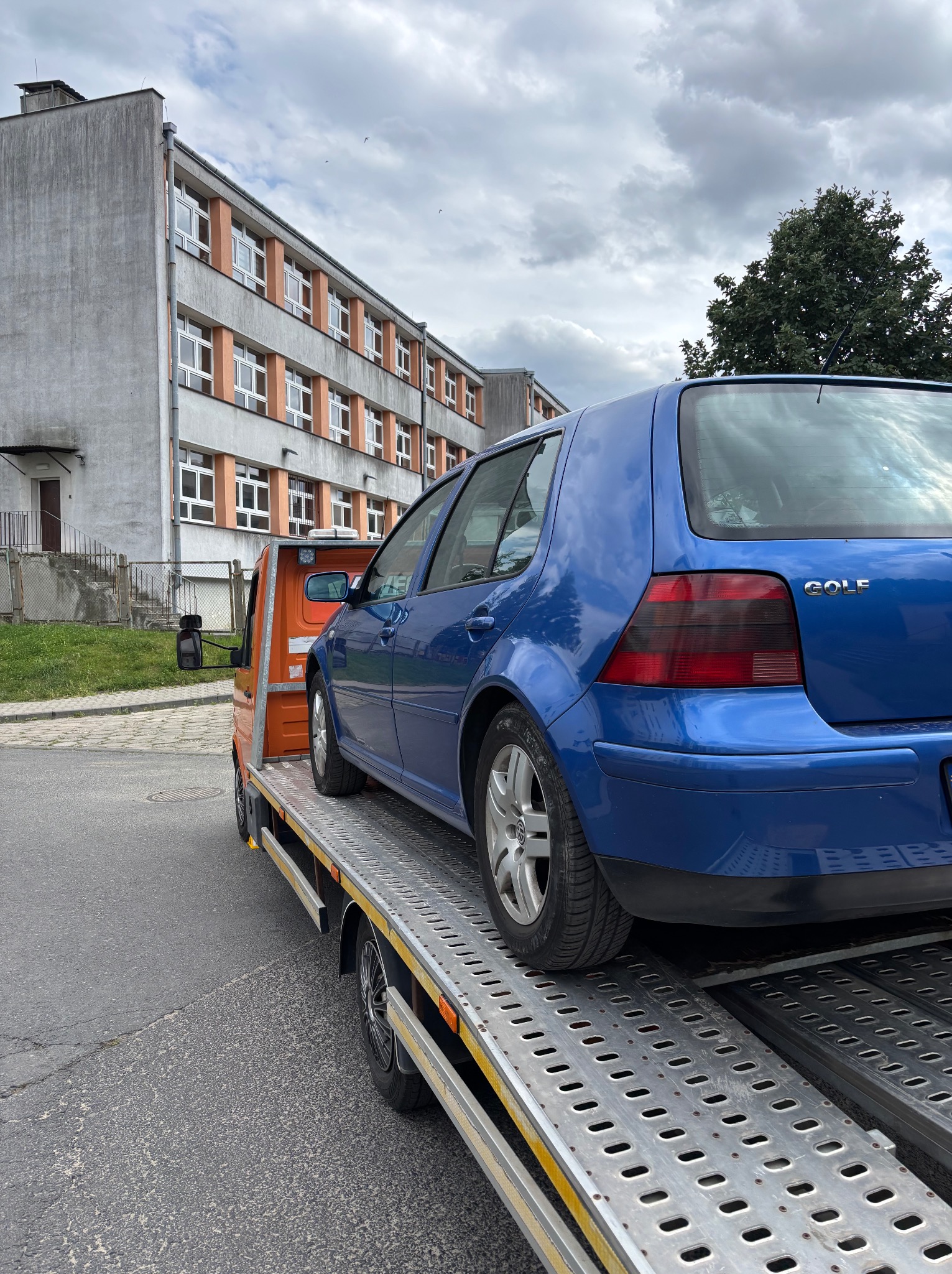 Niebieski Volkswagen Golf na pomarańczowej platformie holowniczej przed budynkiem. Widok z tyłu, pochmurne niebo.
