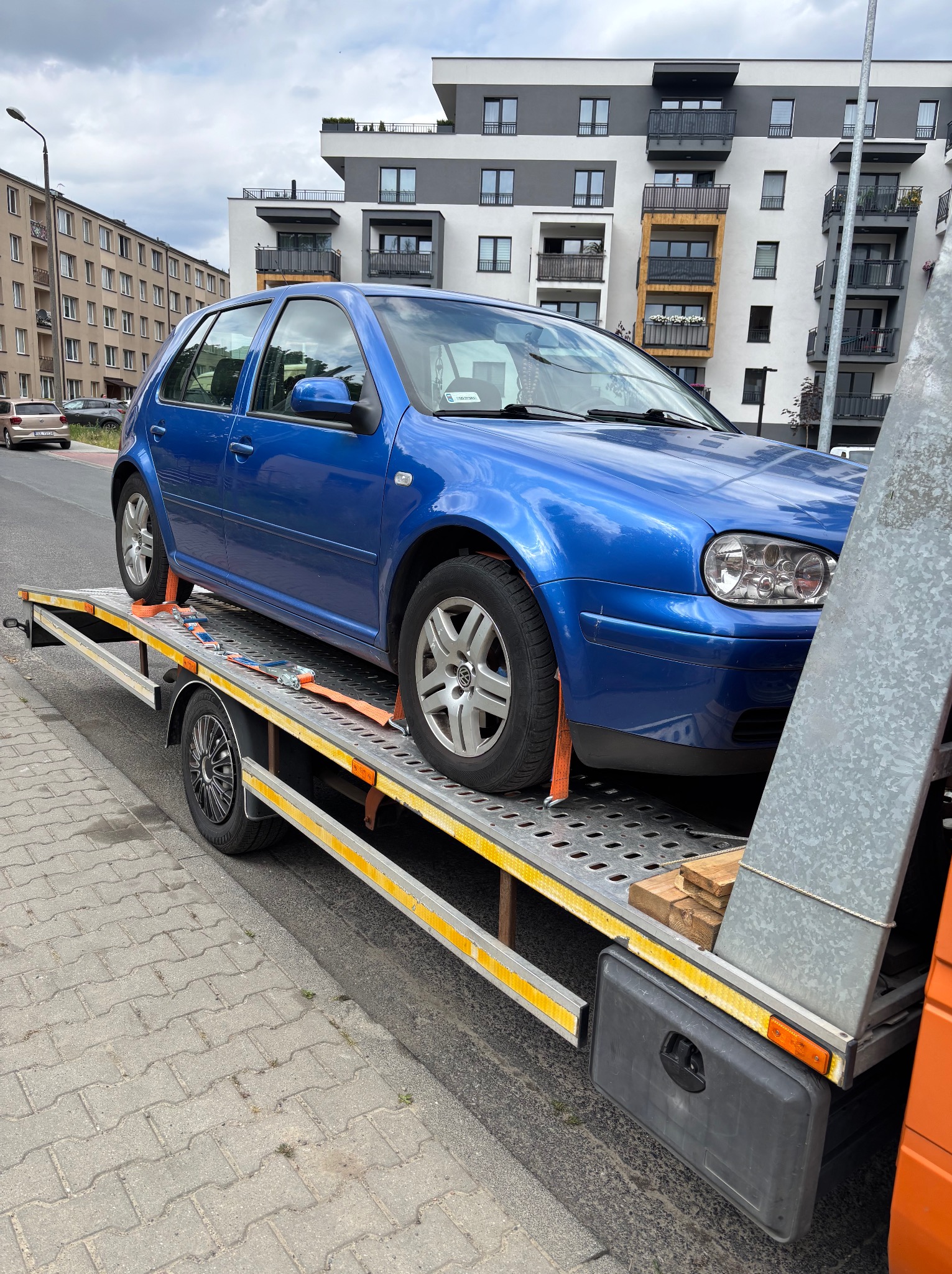 Niebieski Volkswagen Golf zabezpieczony pasami na ażurowej platformie lawety. W tle bloki mieszkalne i chodnik z kostki.