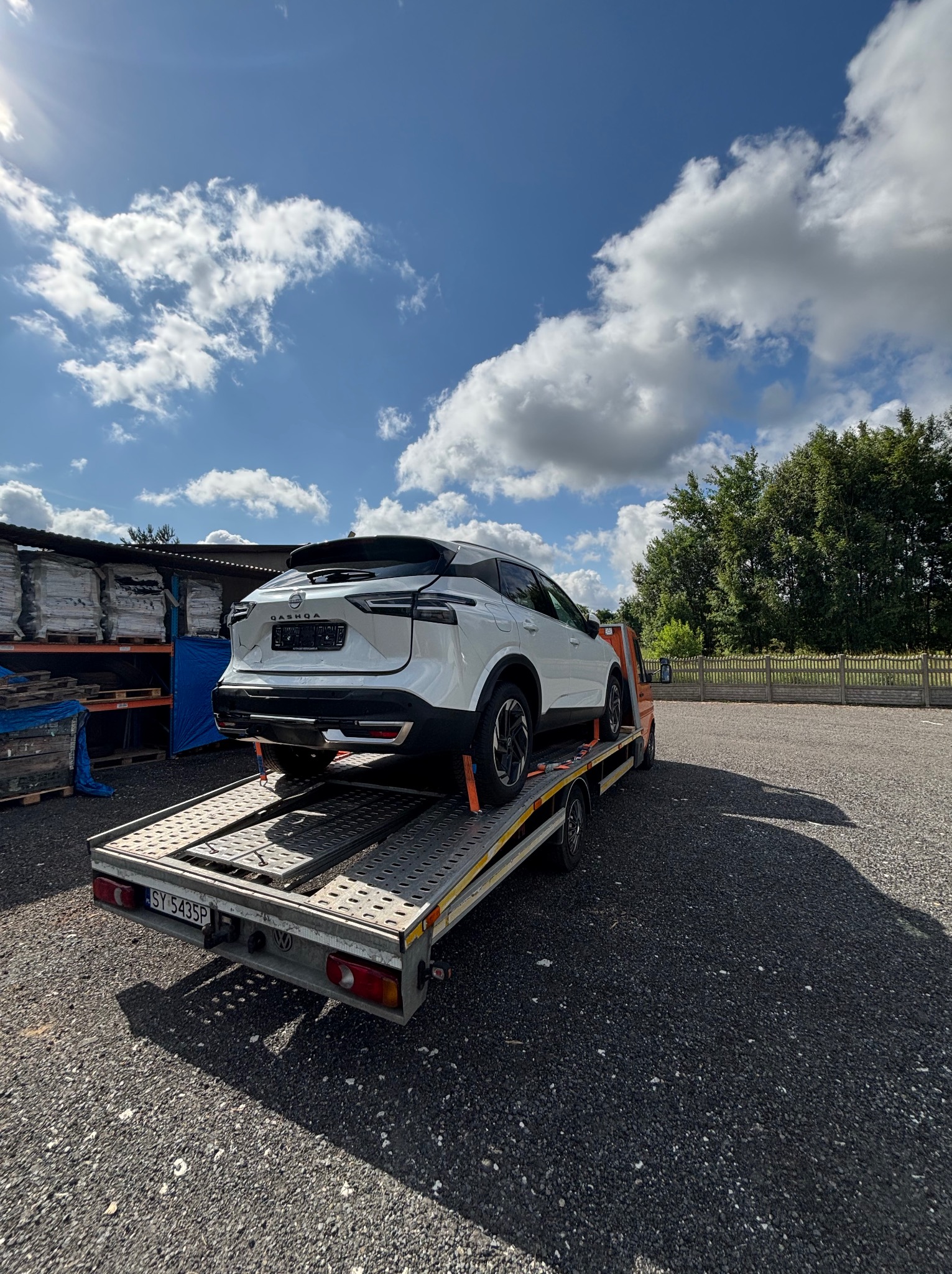 Biały Nissan Qashqai na niskopodwoziowej platformie transportowej w słoneczny dzień. Widoczne tablice rejestracyjne i elementy mocujące pojazd.