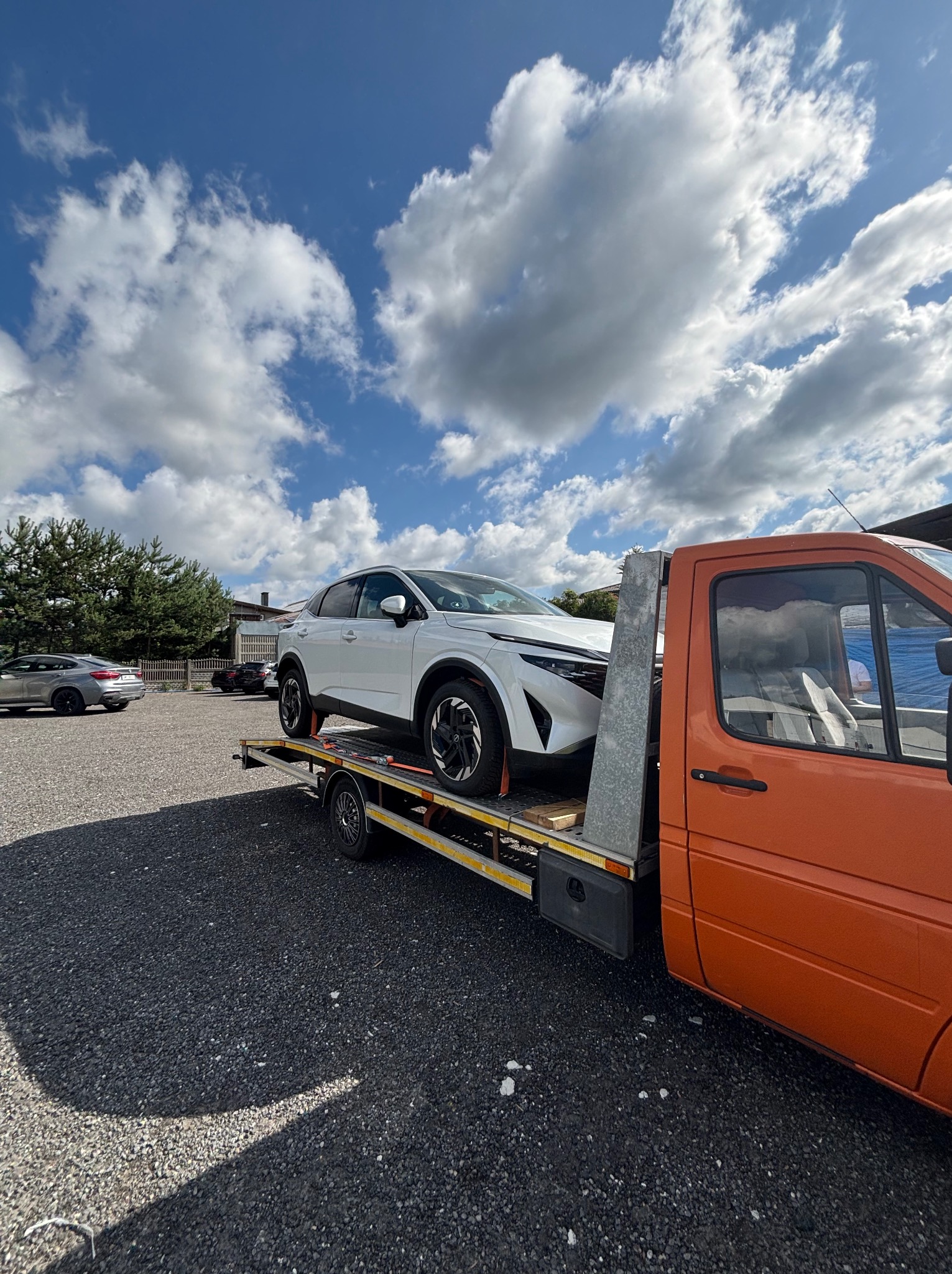 Pomarańczowa laweta z białym samochodem osobowym na platformie, zaparkowana na żwirowym placu pod błękitnym niebem z obłokami.