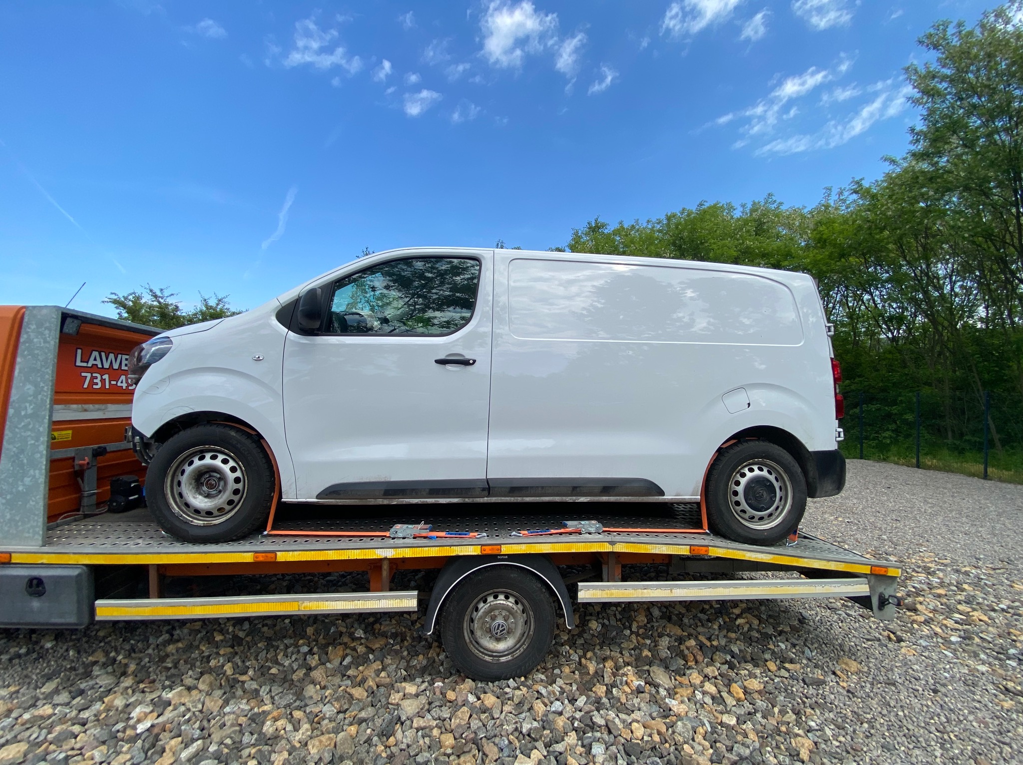 Biały furgon na platformie transportowej z żółtymi elementami zabezpieczającymi, na tle niebieskiego nieba z chmurami i zieleni.