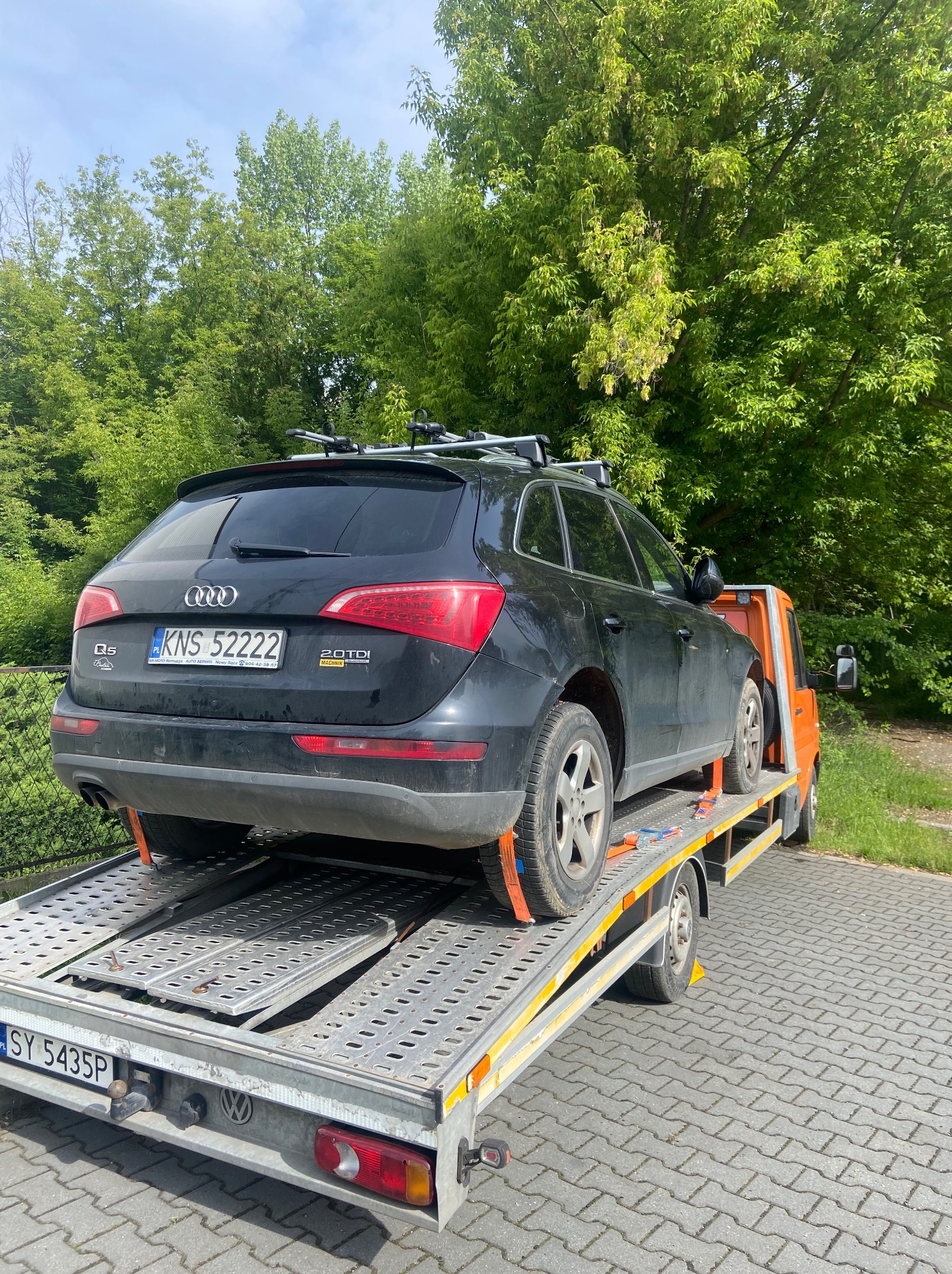 Czarny SUV Audi Q5 zabezpieczony pasami transportowymi na platformie lawety, widok z tyłu na tle zieleni. Rejestracja KNS 52222.