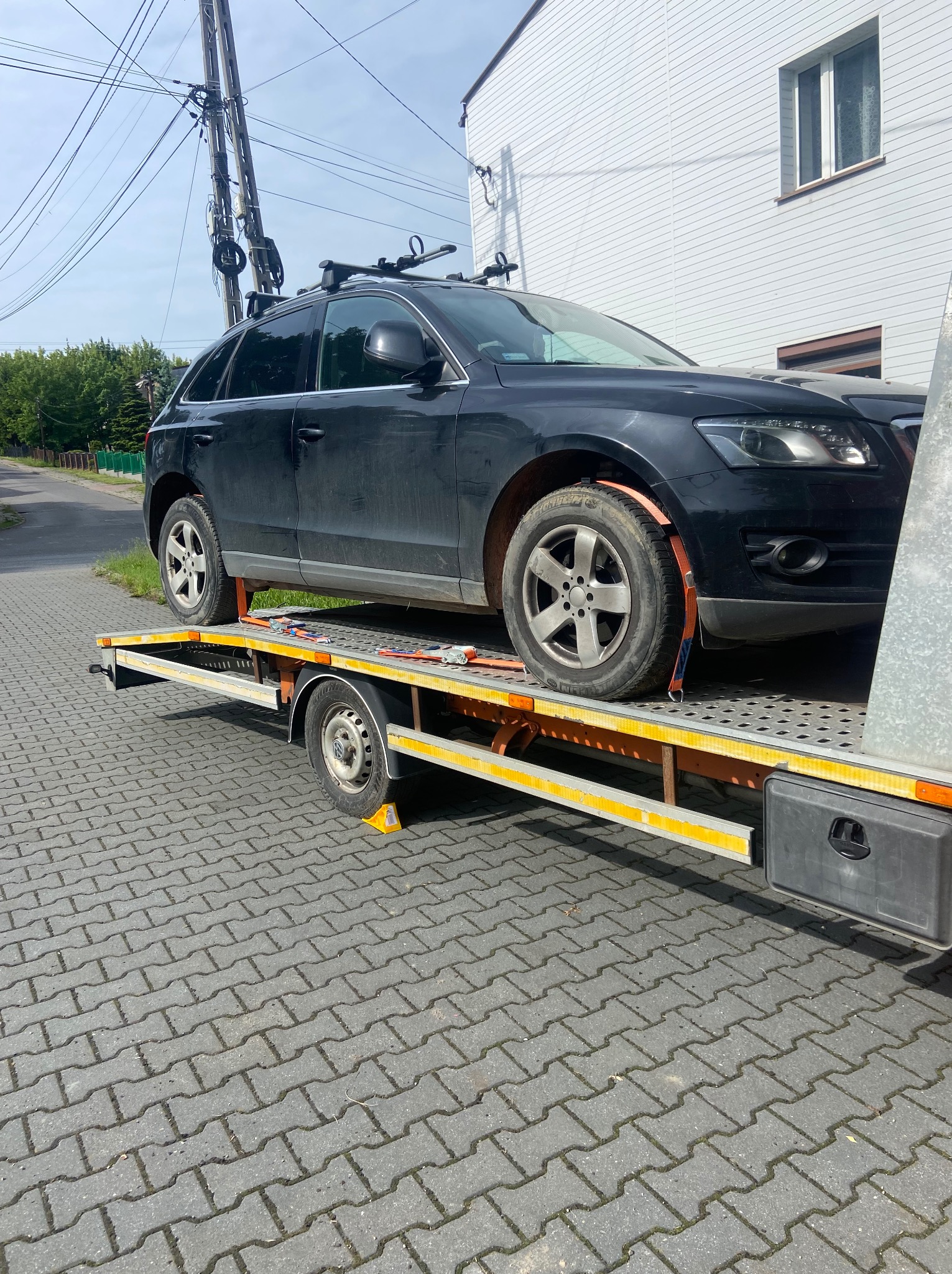 Czarny SUV Audi na żółtej platformie transportowej, zabezpieczony pasami, na tle brukowanej drogi i budynku z białą elewacją.