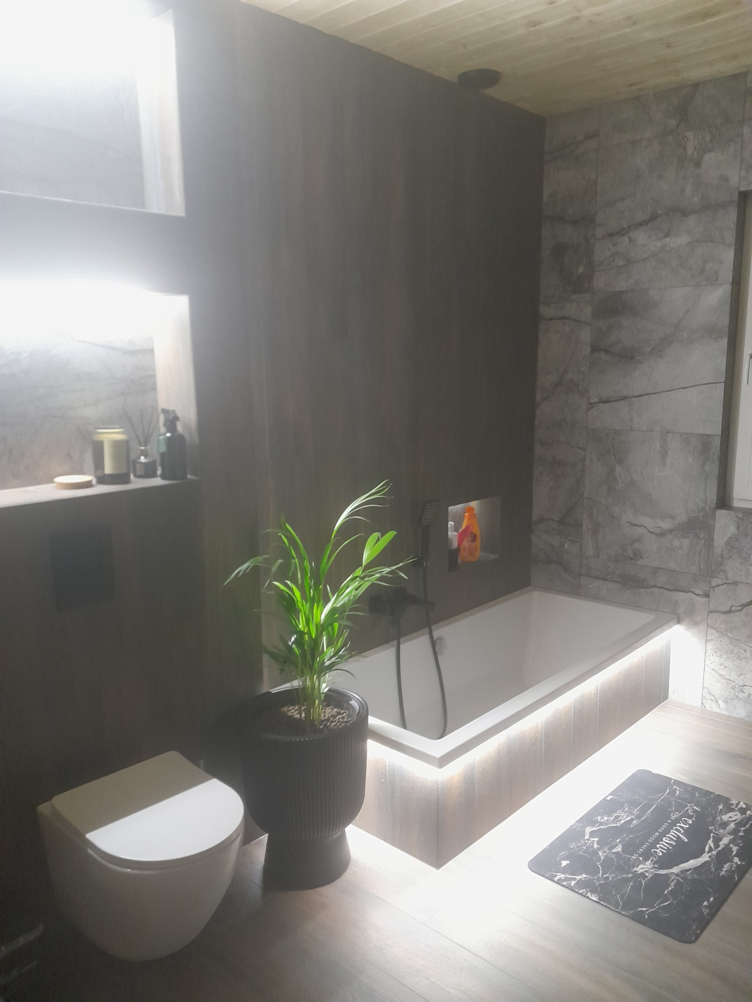 Nowoczesna łazienka z ciemnymi panelami ściennymi, podświetloną wanną, wiszącą toaletą i rośliną doniczkową w czarnej donicy.