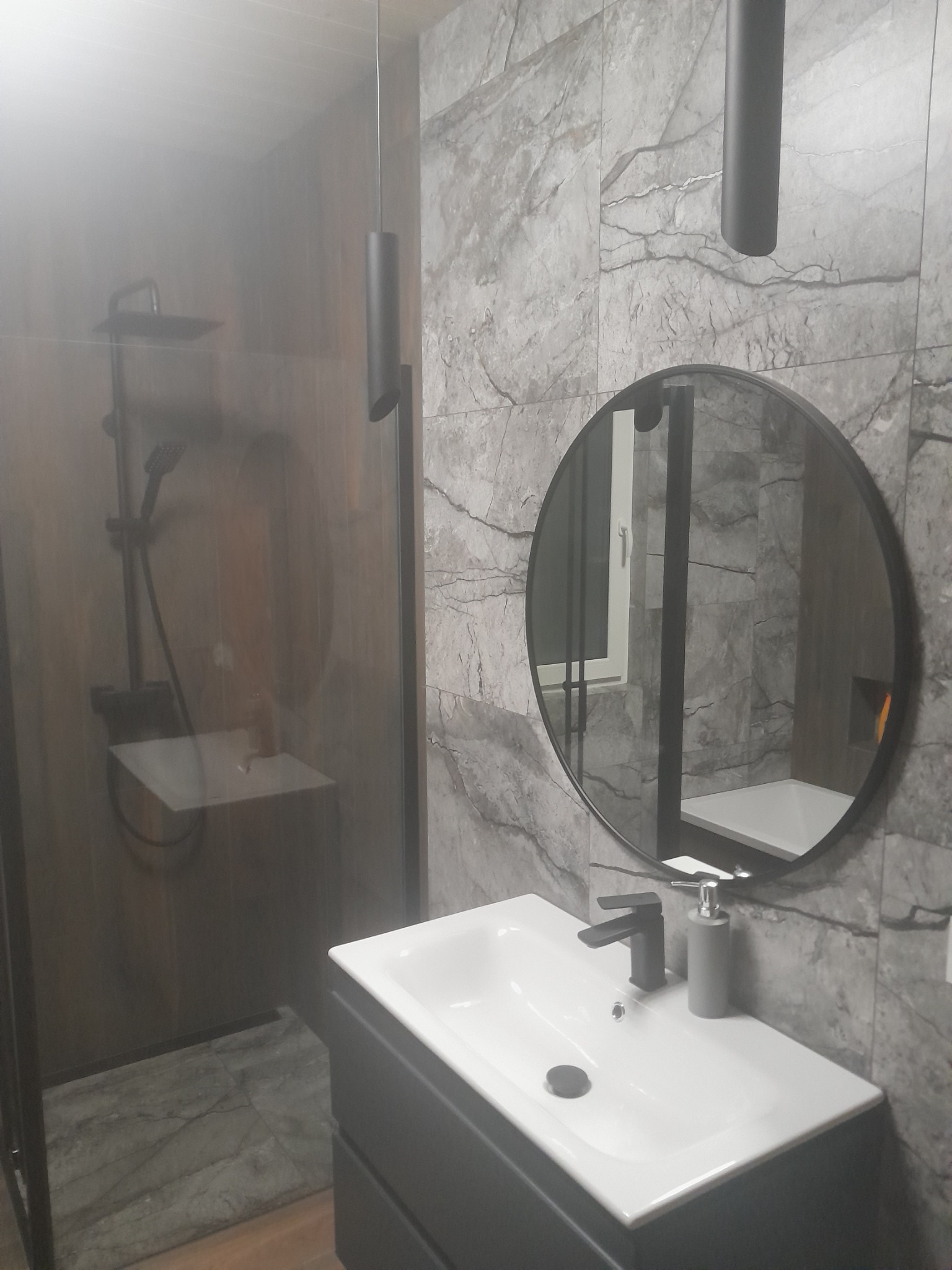 Nowoczesna łazienka z kabiną prysznicową typu walk-in, umywalką na szafce i okrągłym lustrem na tle ściany wyłożonej płytkami imitującymi marmur; czarne akcenty w postaci armatury i oświetlenia.