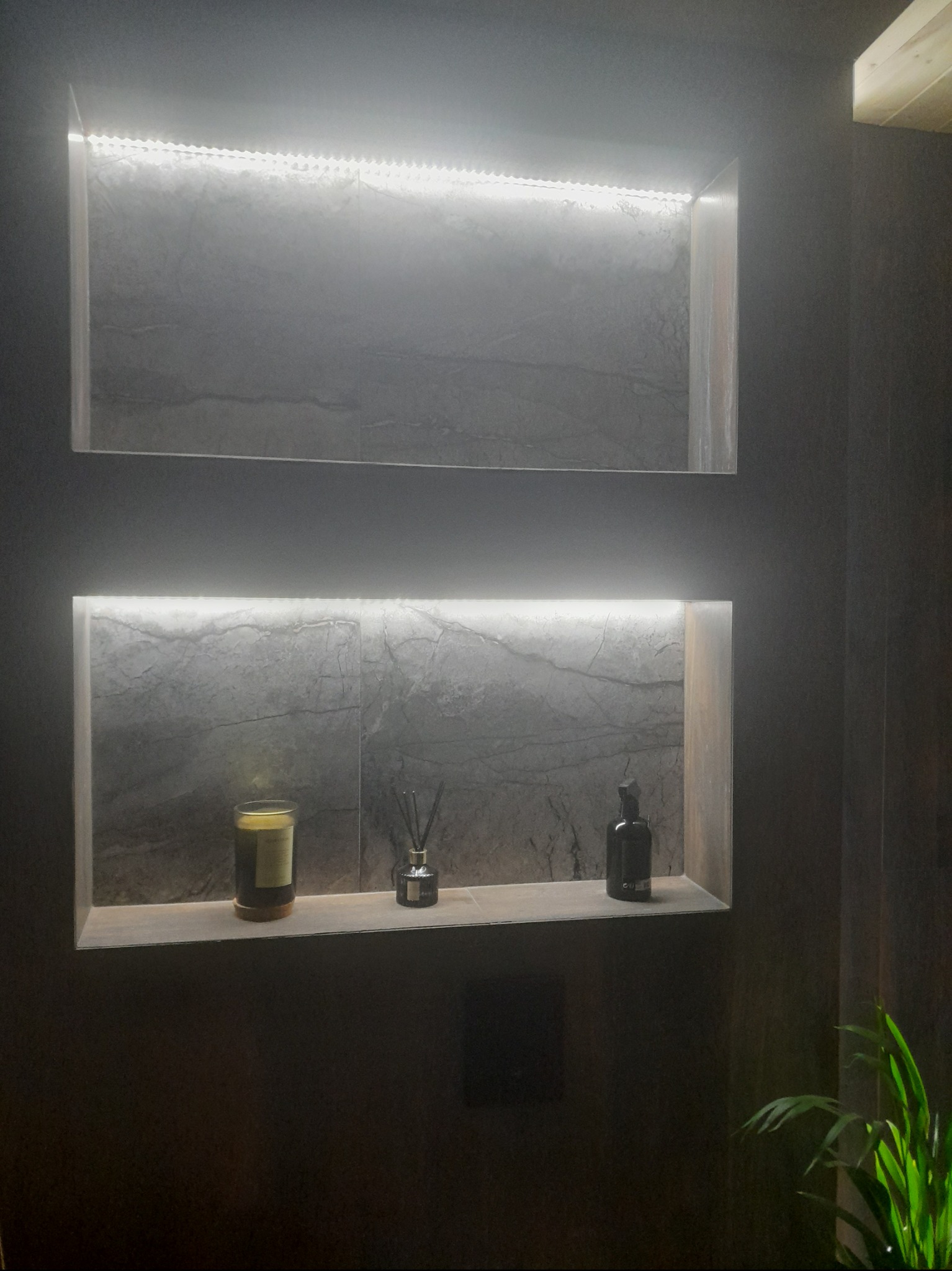 Dwa podświetlane wnęki w ciemnej łazience, wyłożone płytkami imitującymi marmur, z dekoracjami: świecą zapachową, dyfuzorem zapachowym i butelką z dozownikiem.