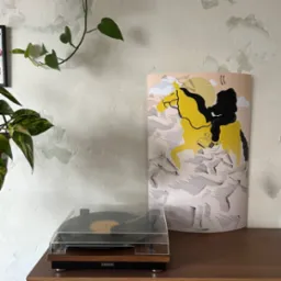 Plakat z ilustracją jeźdźca na żółtym koniu w sportowych butach, stojący na drewnianej komodzie obok gramofonu Lenco, na tle ściany z dekoracyjną fakturą i roślin doniczkowych.