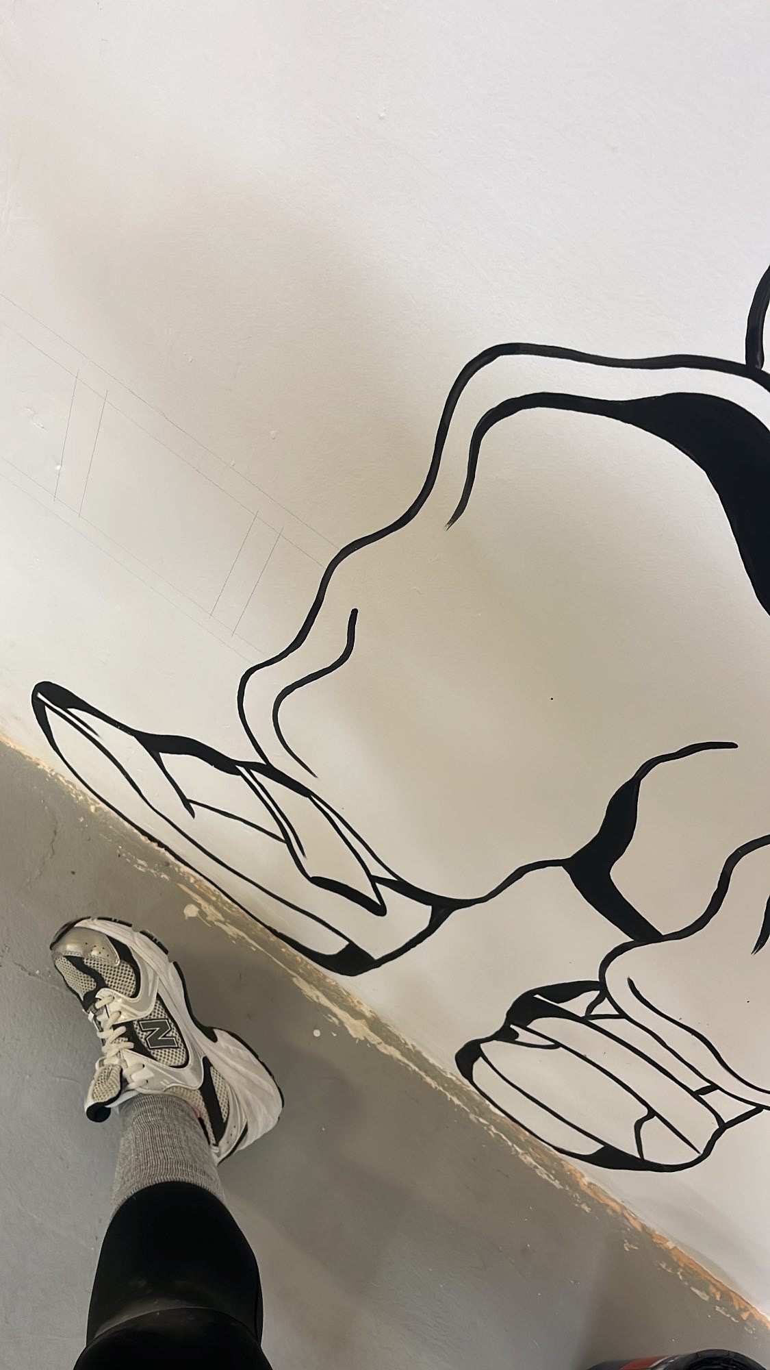 Czarno-biały mural przedstawiający dłonie na białej ścianie, widoczna noga w sportowym bucie i szarej skarpetce, ujęcie z dołu.