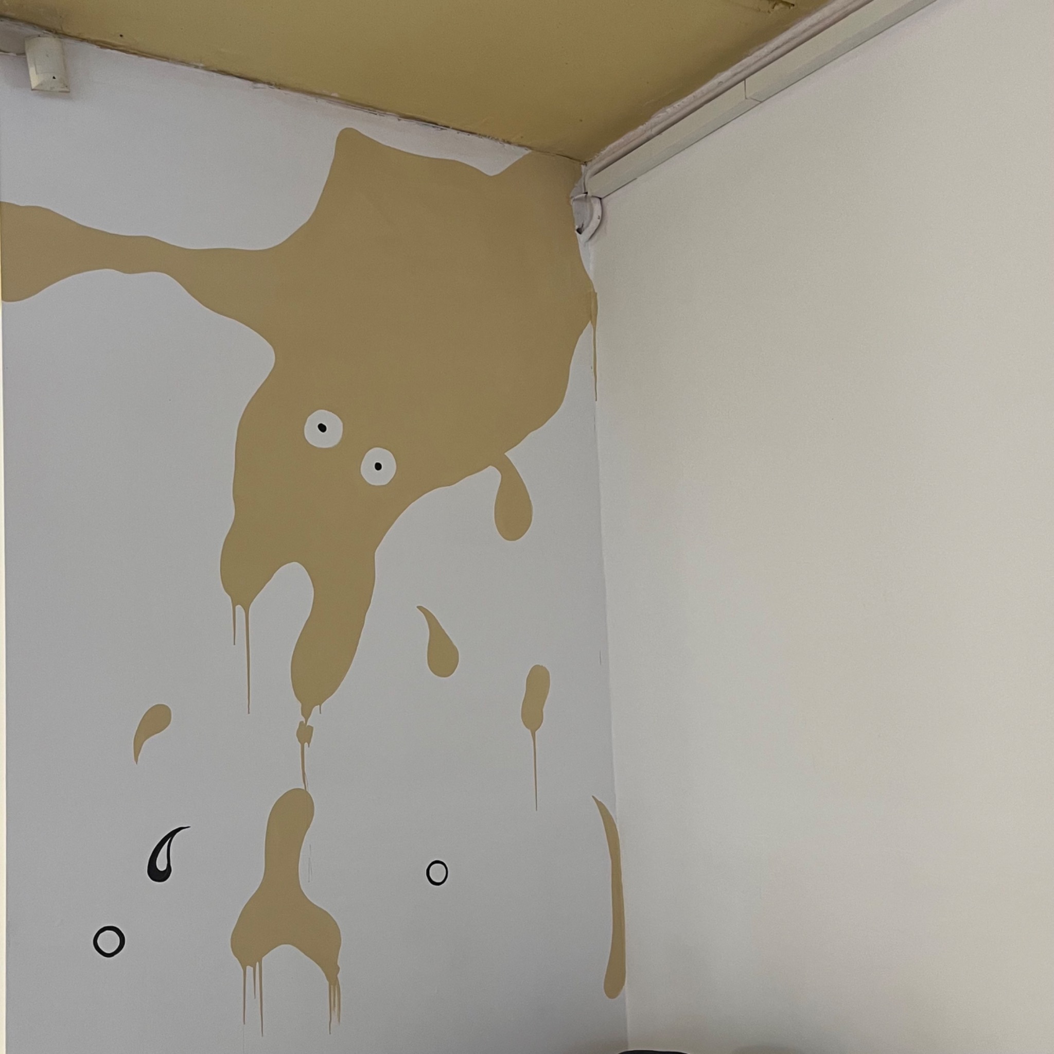 Abstrakcyjna malatura ściany w odcieniach beżu i czerni, przypominająca rozlaną plamę z dwoma okrągłymi oczami, w rogu pomieszczenia.